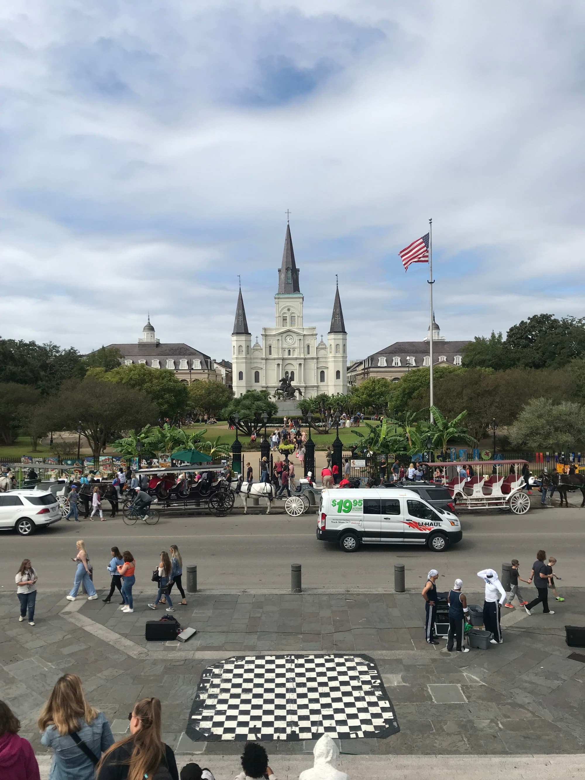 Jackson Square in New Orleans with people walking, vehicles and carriages on the street and a white historic building in view.