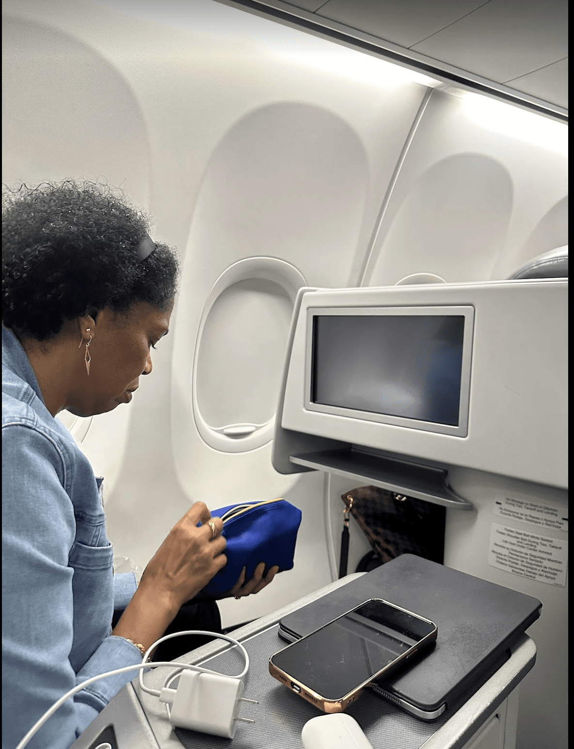 A person sitting in a chair on an airplane with a screen in front of them. They are looking down at their hands, which are holding a cobalt blue bag. There is a phone on the table next to them with a charger.