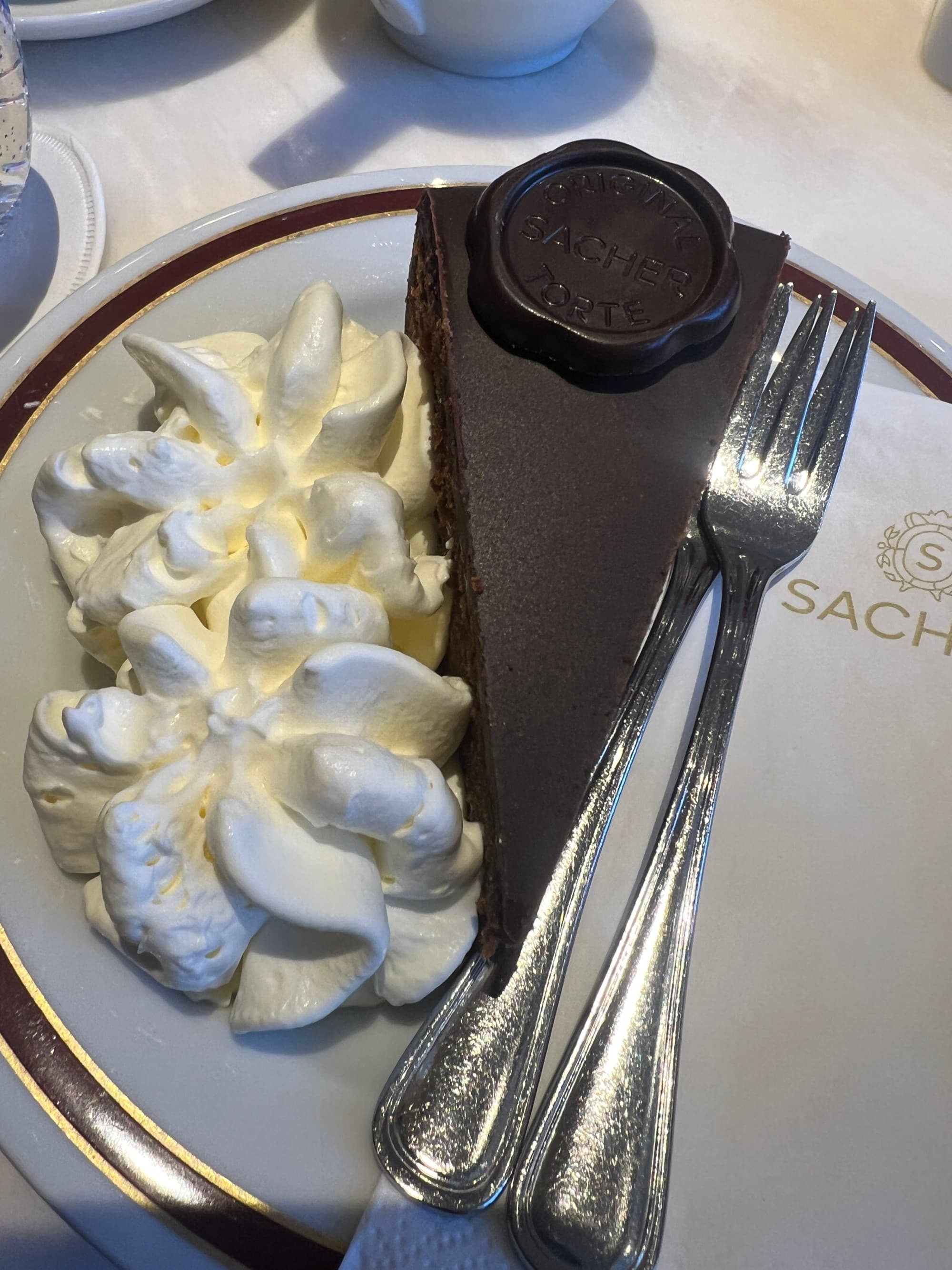 An image of a slice of chocolate cake with a floral shaped chocolate design on top of it and a pile of whipped cream to the left of the slice. There is silver flatware on the right side, all positioned atop of a white plate.