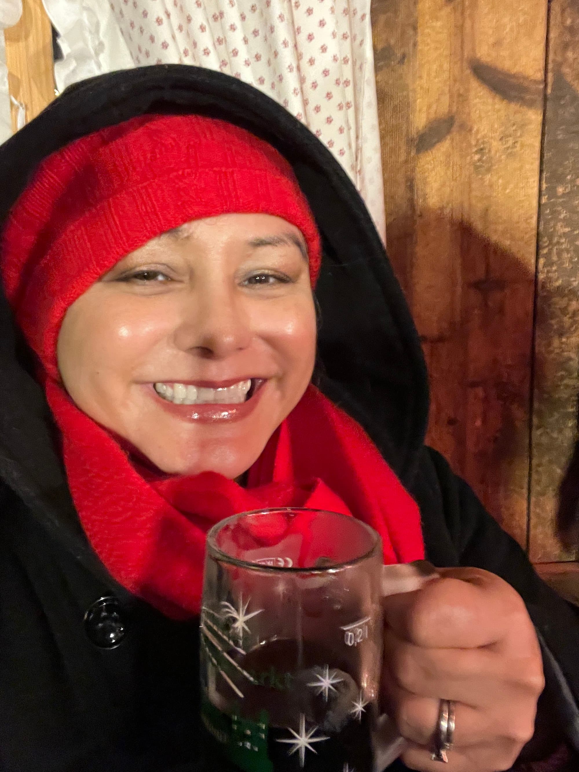 An image of a person wearing a red knit hat and a black hoodie smiling for a selfie while holding a glass mug against a wooden wall.