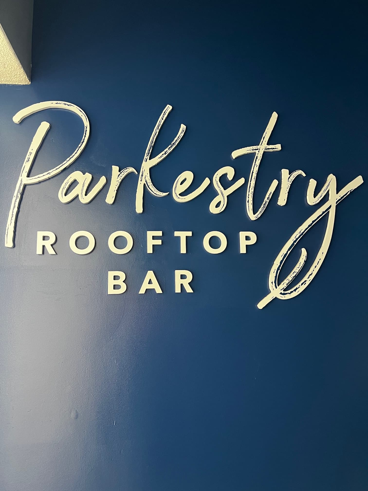 This image depicts a blue wall with white signage attached to it that reads "Parkestry Rooftop Bar".