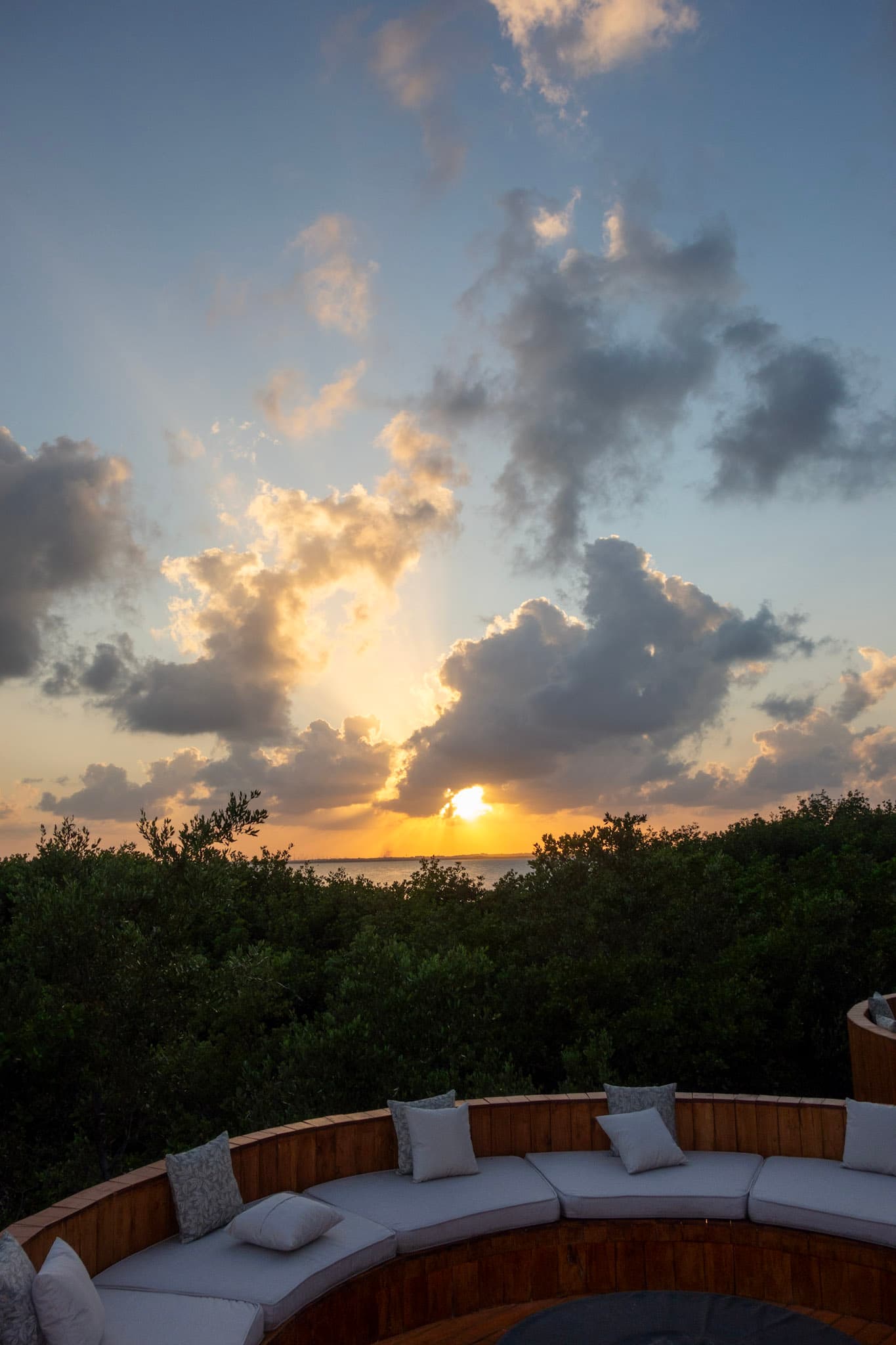 An outdoor lounge area of the hotel bordered by green bushes, with the sea just beyond at sunset.
