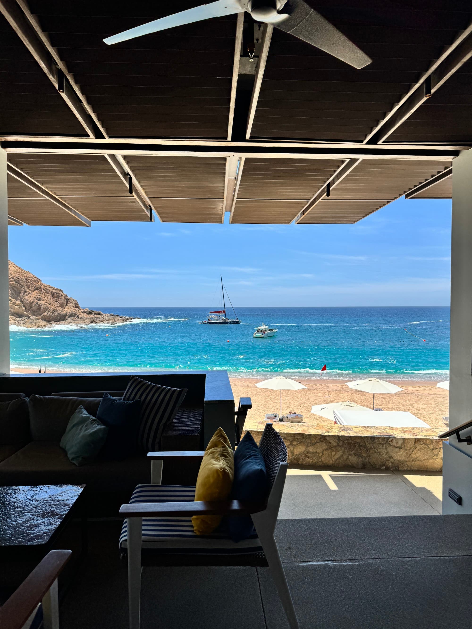 A view of the resort's beach, from a covered lounge area, showcasing the bright blue waters, sandy shoreline, and a couple of small boats on a sunny day.