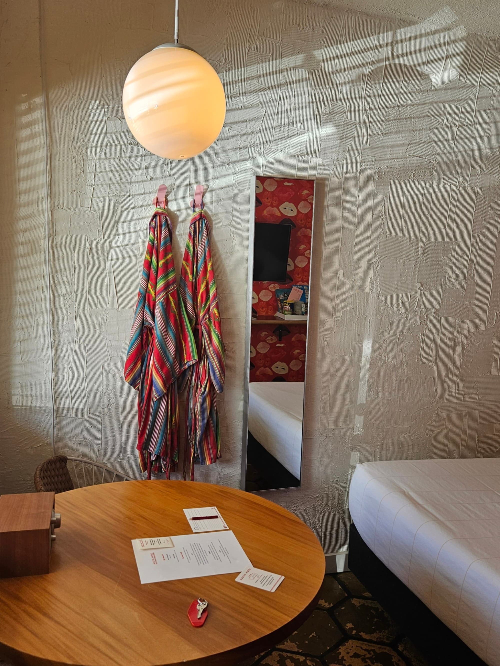 The interior of a motel room, with a round brown table, hanging light fixture and two colorful robes on the wall - Nicole Diez