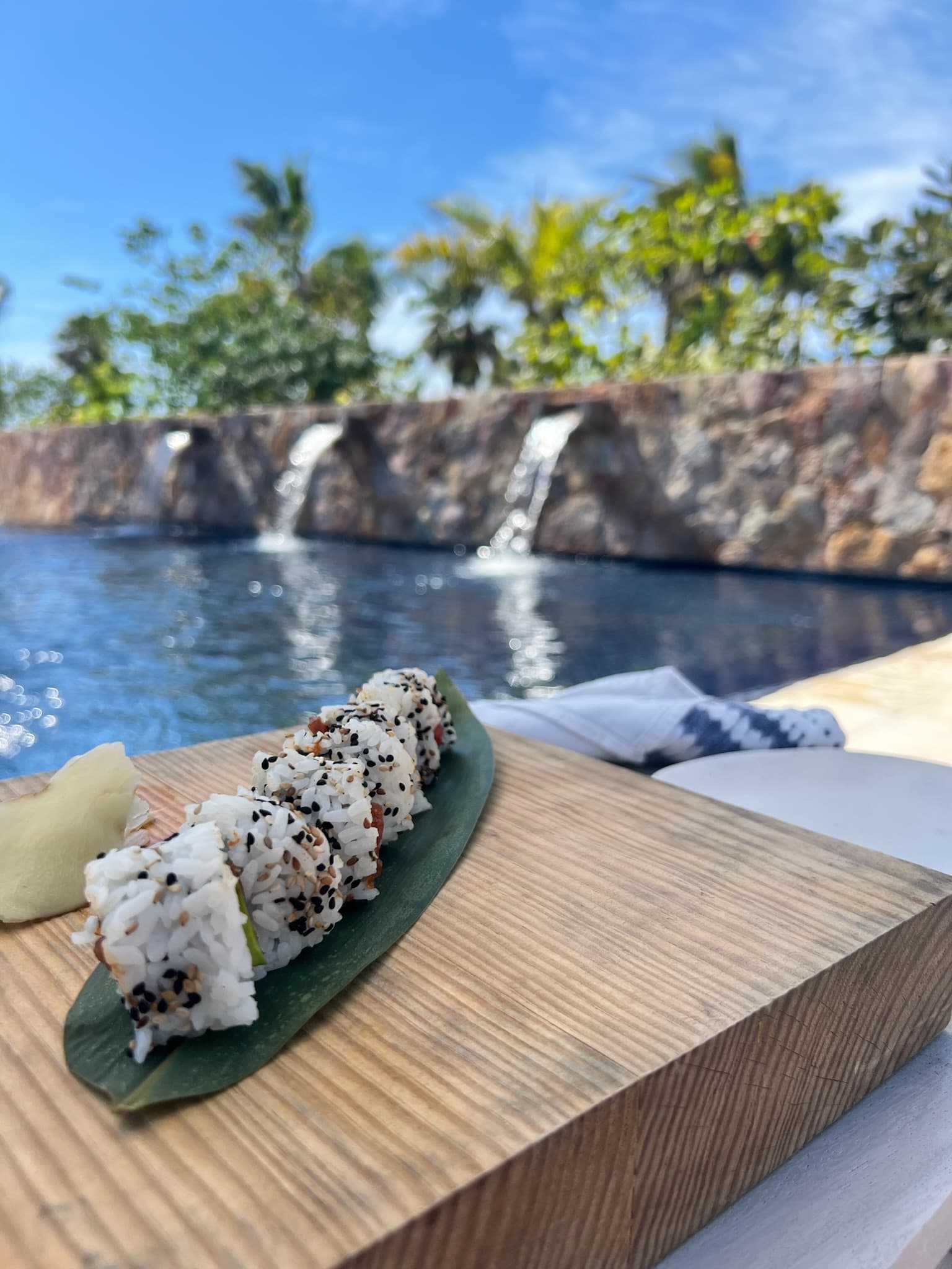 A freshly prepared sushi roll, plated on an outdoor table in the afternoon, with a pool and tropical trees in the background.