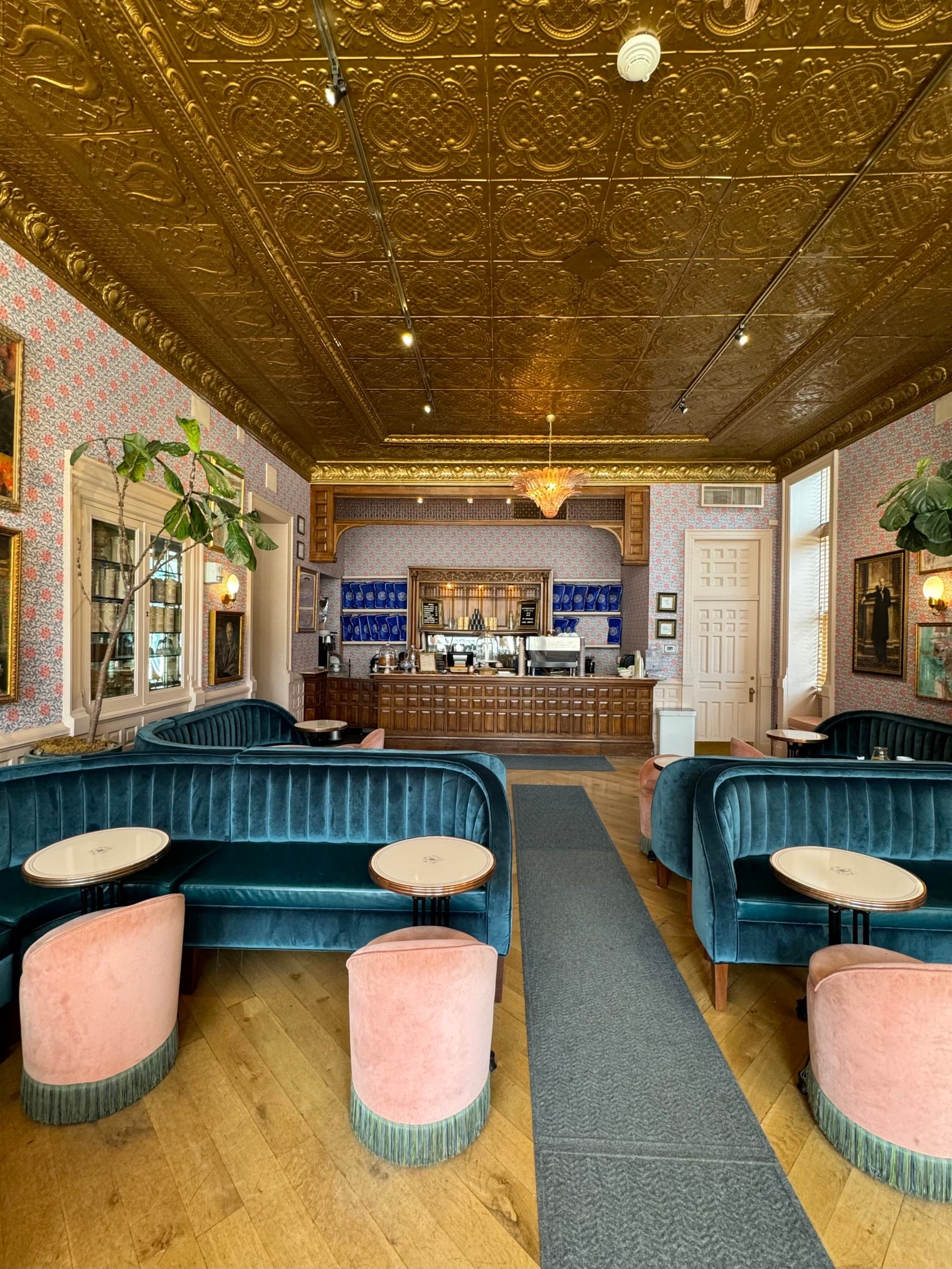 One of the hotel's dining areas, with a gold, gilded ceiling, small round tables, blue couches, salmon-colored stools and a bar.