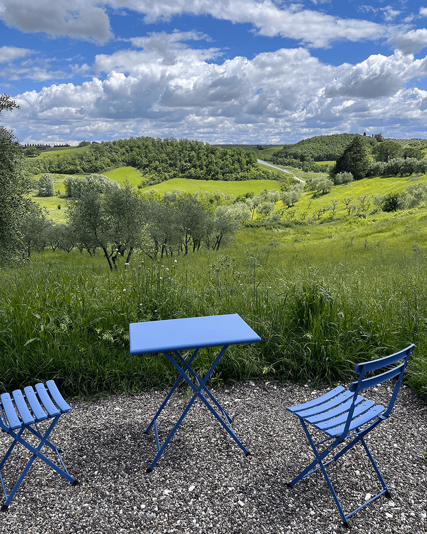 Blue chairs and table set in the Tuscan landscape, with a view of the green fields and blue sky in the distance.