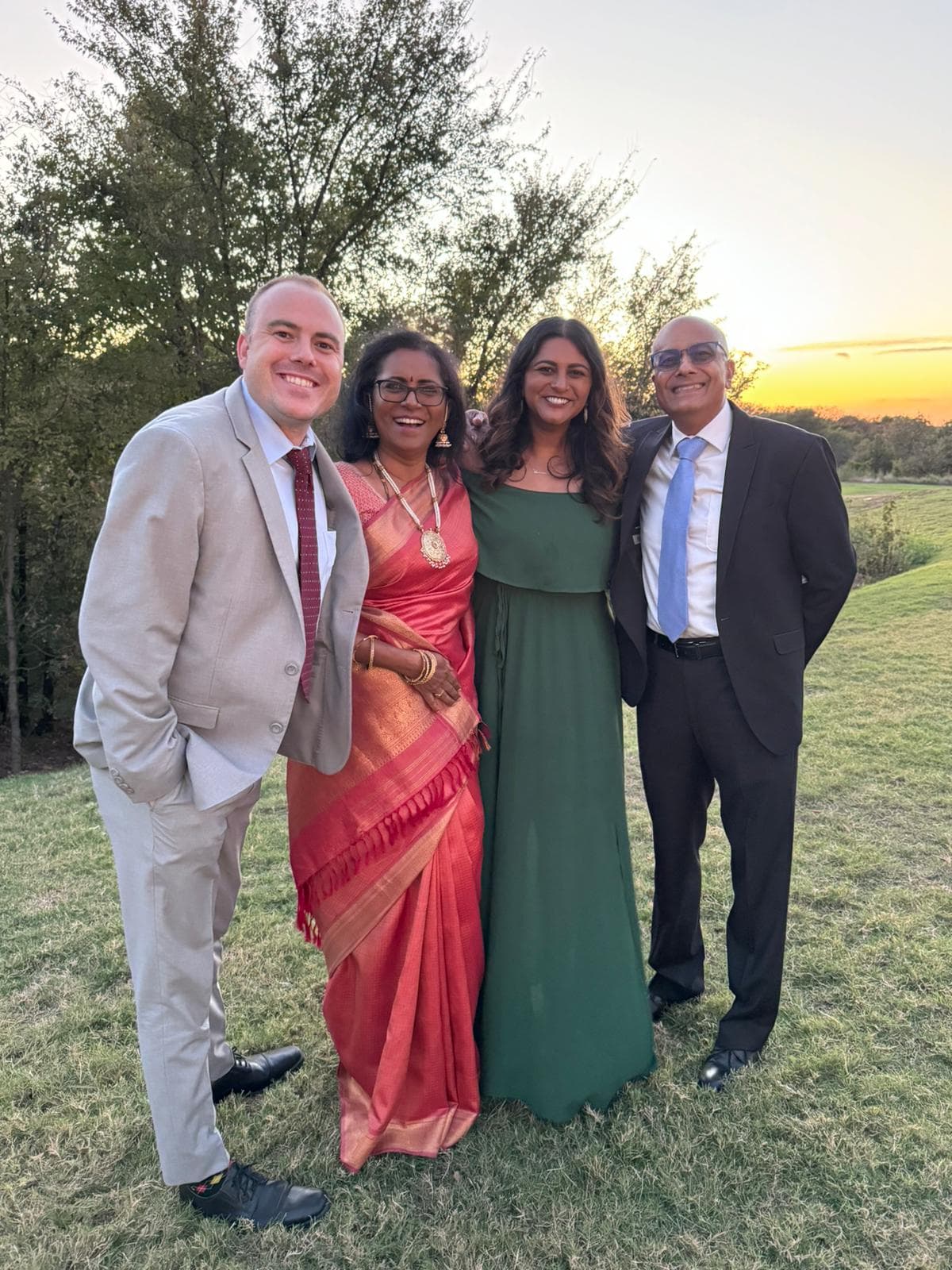 two men and two women in formal clothing standing on grass with sunset beyond
