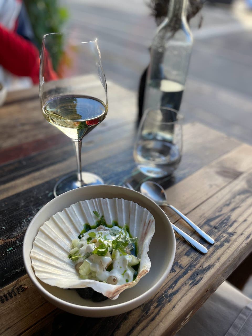 A wooden table with an inventive dish of a filled clam shell next to a glass of wine - Leah Chen