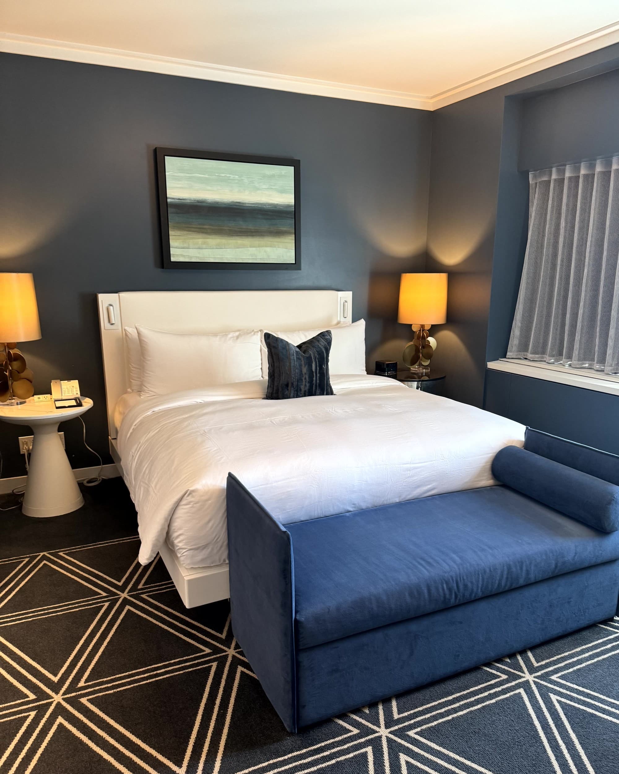 A view of a hotel room with a queen size bed, lamps, a navy blue bench and triangle printed navy and cream carpet. Accommodations - Fairmont Gold 1 - Rondene Grinam