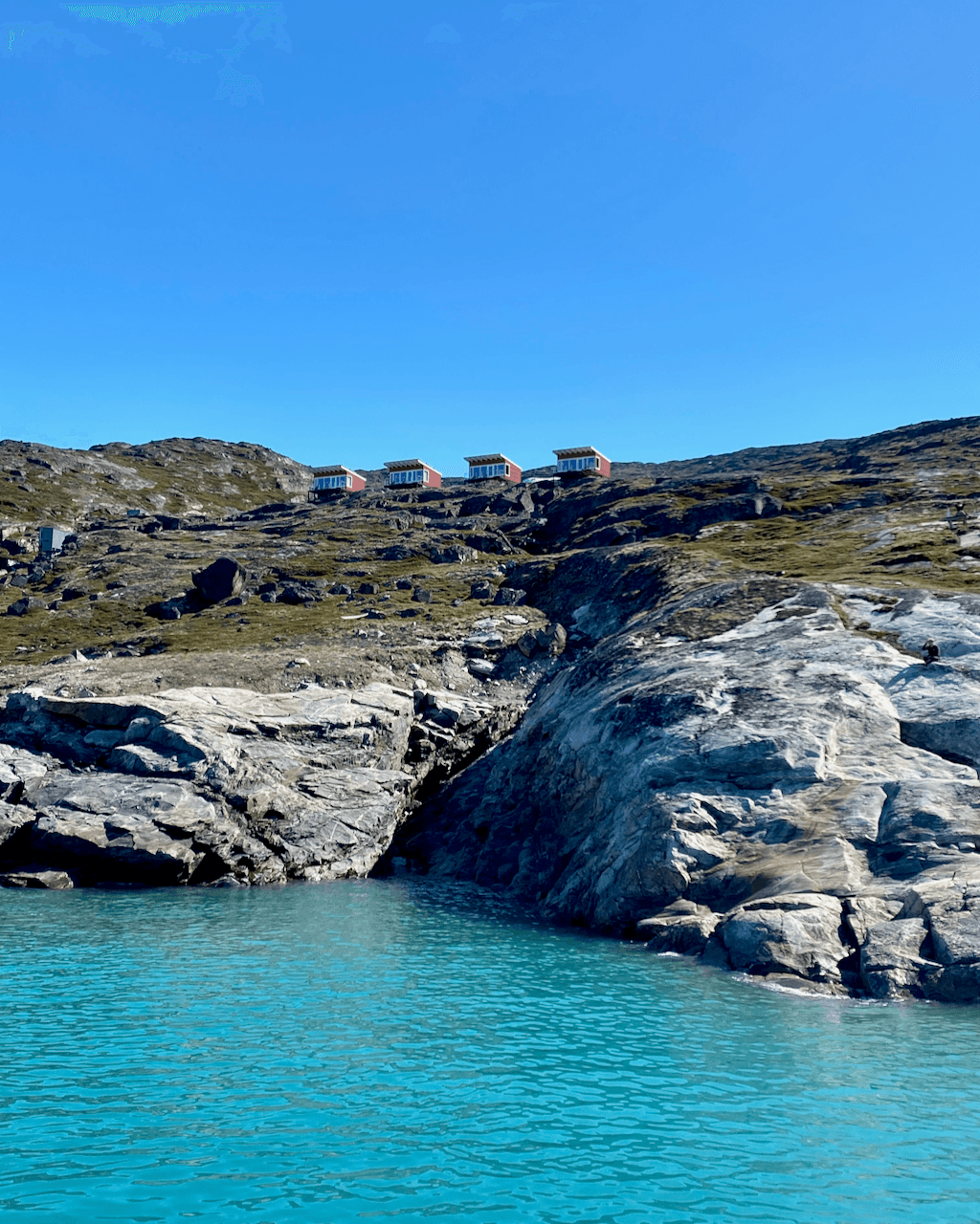 A view of four cabins on the side of a hill overlooking rocky shoreline and bright turquoise water.