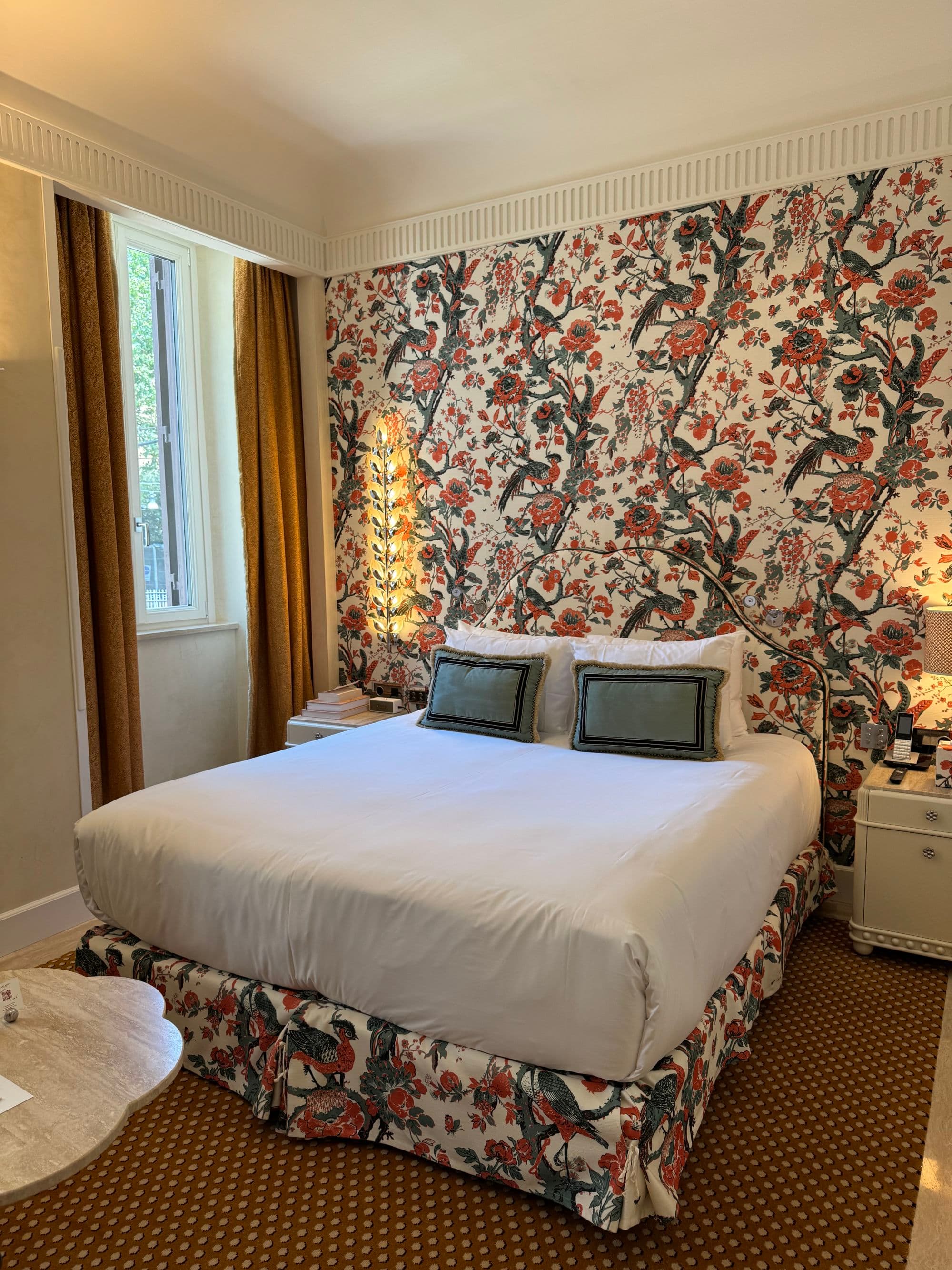 The hotel bedroom has bright wallpaper and bed coverings and quaint furnishings.