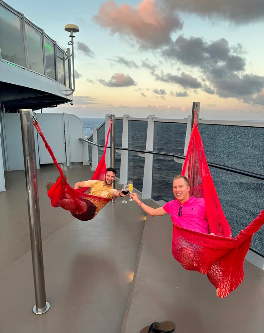 A picture of two guys hanging in red hammocks and cheersing with the water and sunset behind them.