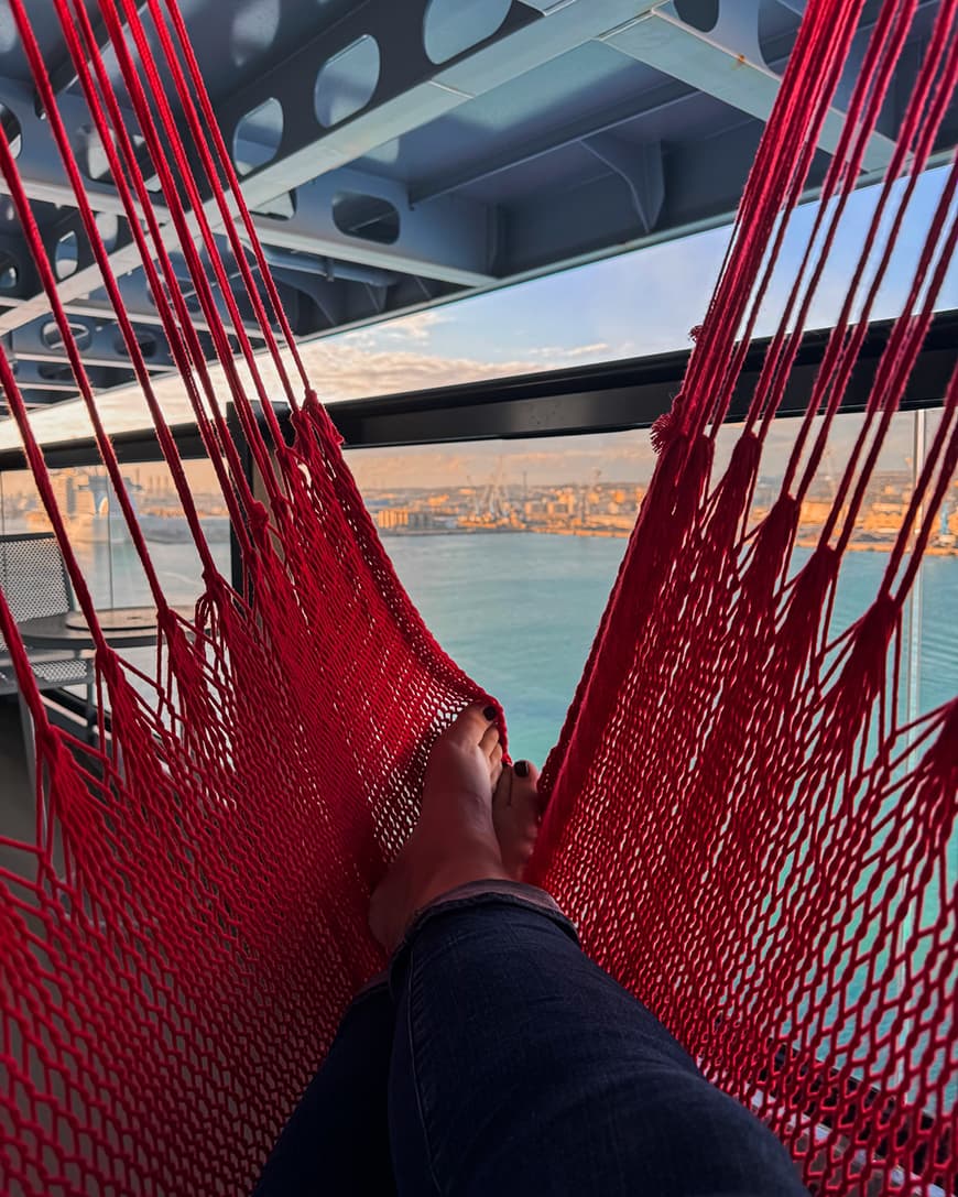 A person's feet laying on a red hammock with a slight view of a body of water in the background