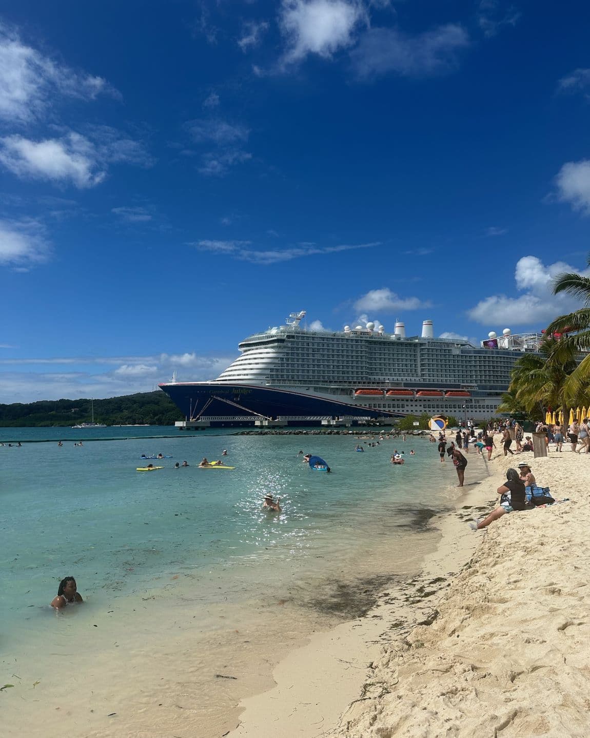 A view of the Carnival Jubilee Honduras along the coastline with people scattered all over the beach and swimming in the ocean.