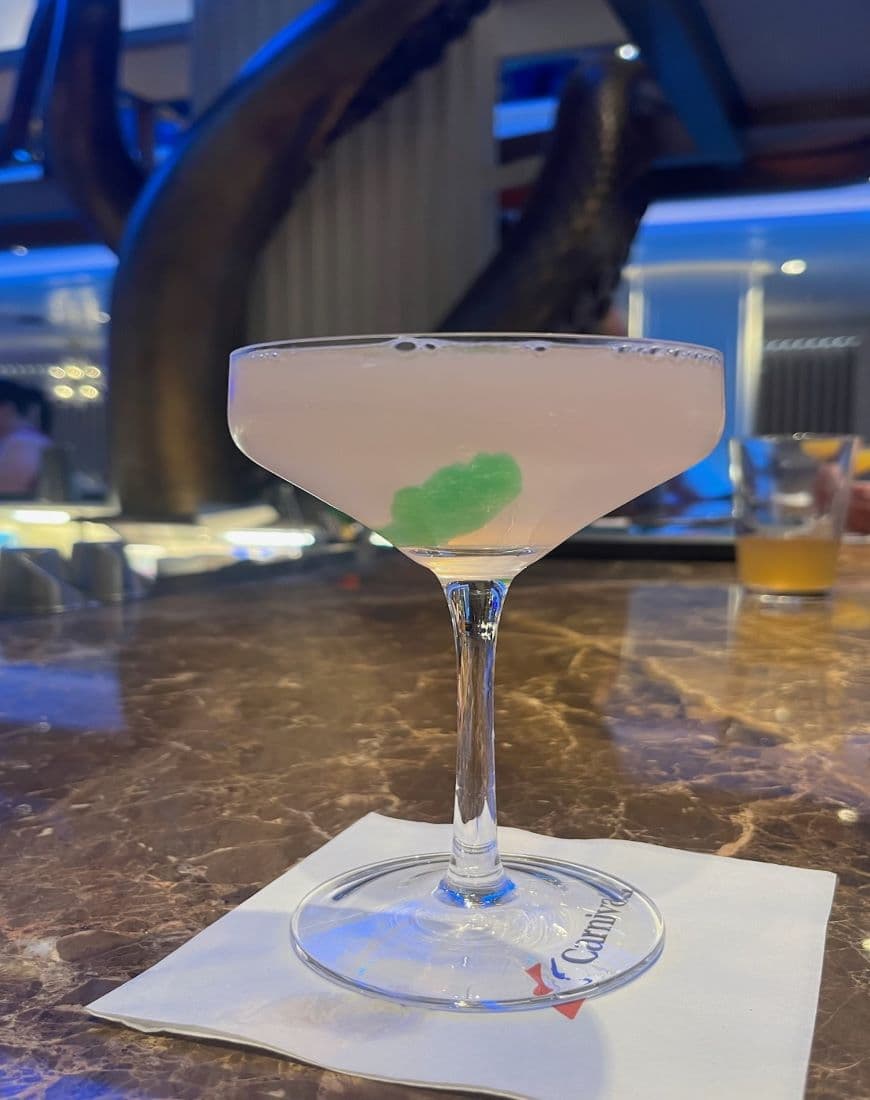 A pink cocktail with a green garnish on a bar napkin on a counter.