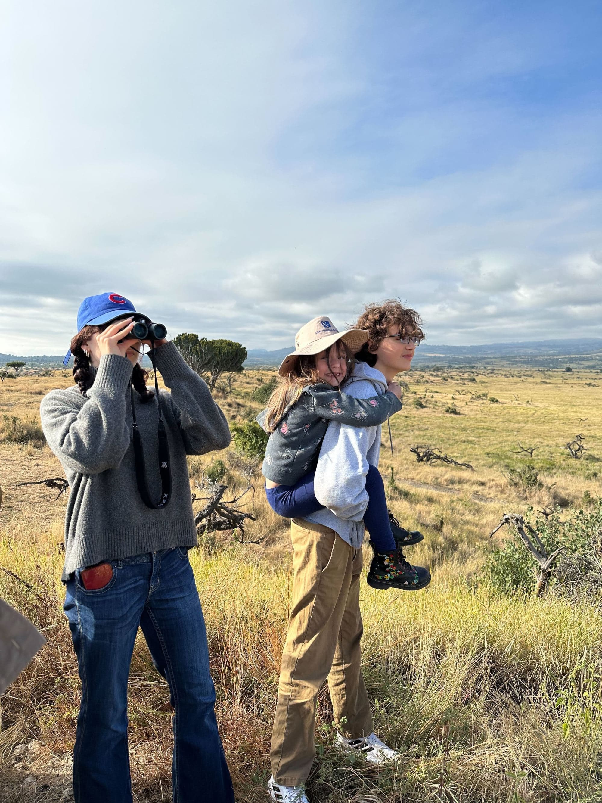 A young woman looks through binoculars and stands near a young man with his little sister piggy backing him