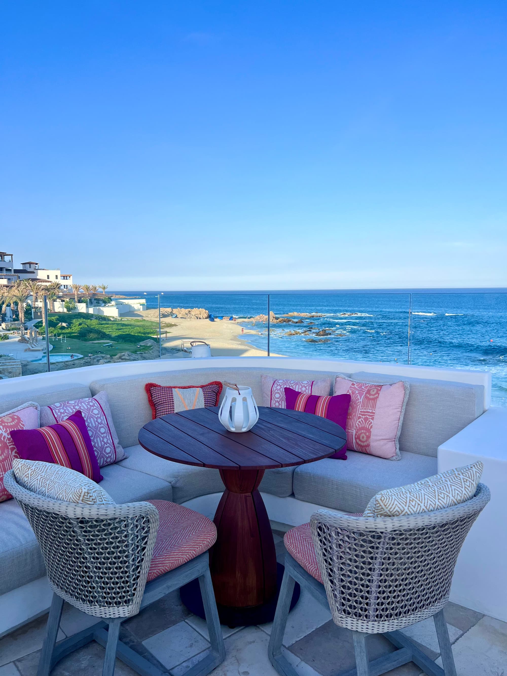 A white corner sofa with pink cushions and two chairs overlooking the ocean at Sora Rooftop Bar on a sunny day.