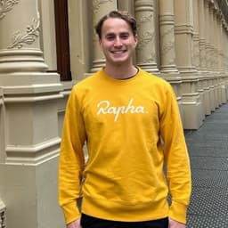 a man in a yellow crew-neck sweater smiles outdoors
