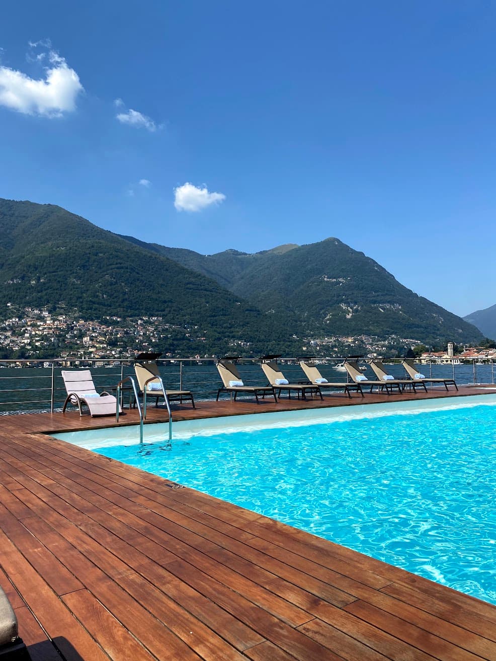 View of a bright blue pool with wooden deck and empty lounges to one side in a sunny costal area with mountains in the background