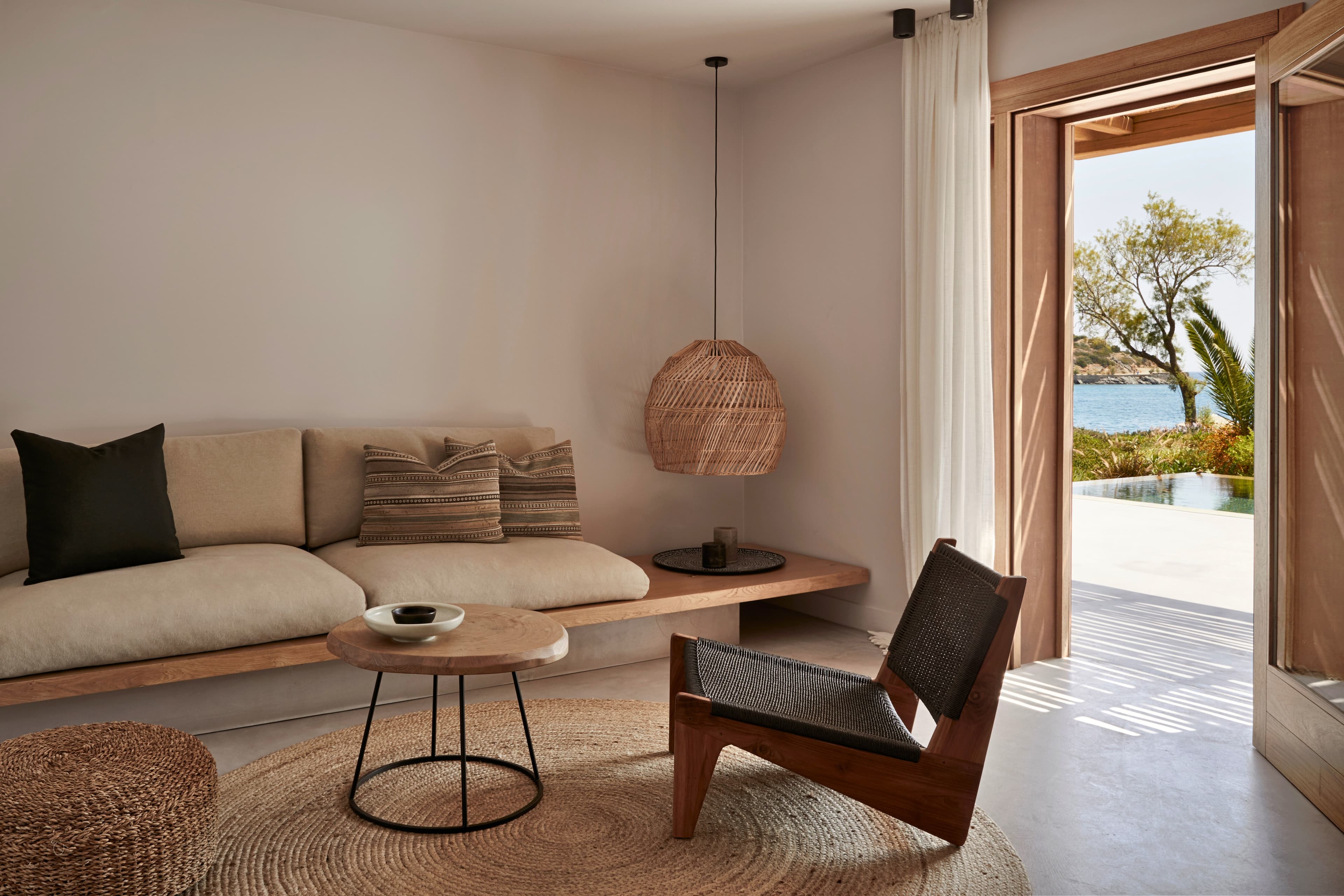 A living area of Minos Beach Art Hotel with neutral shades of beige and wood, with boho chic style and an open doorway leading out to a body of water.