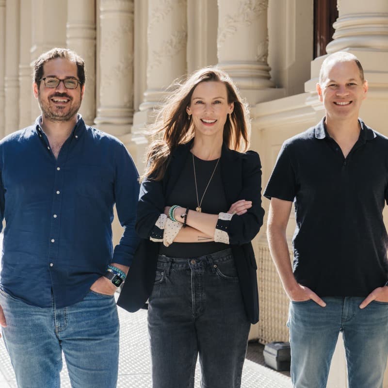 two men and a woman smile together outside on a sunny day