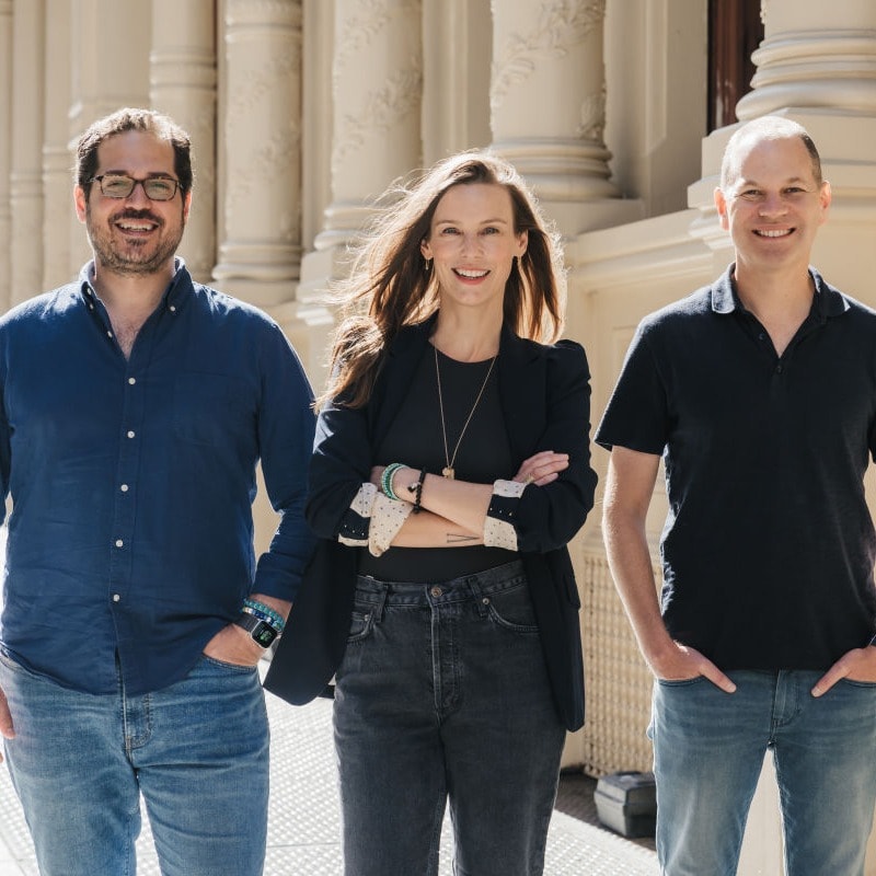 two men and a woman smile together outside on a sunny day