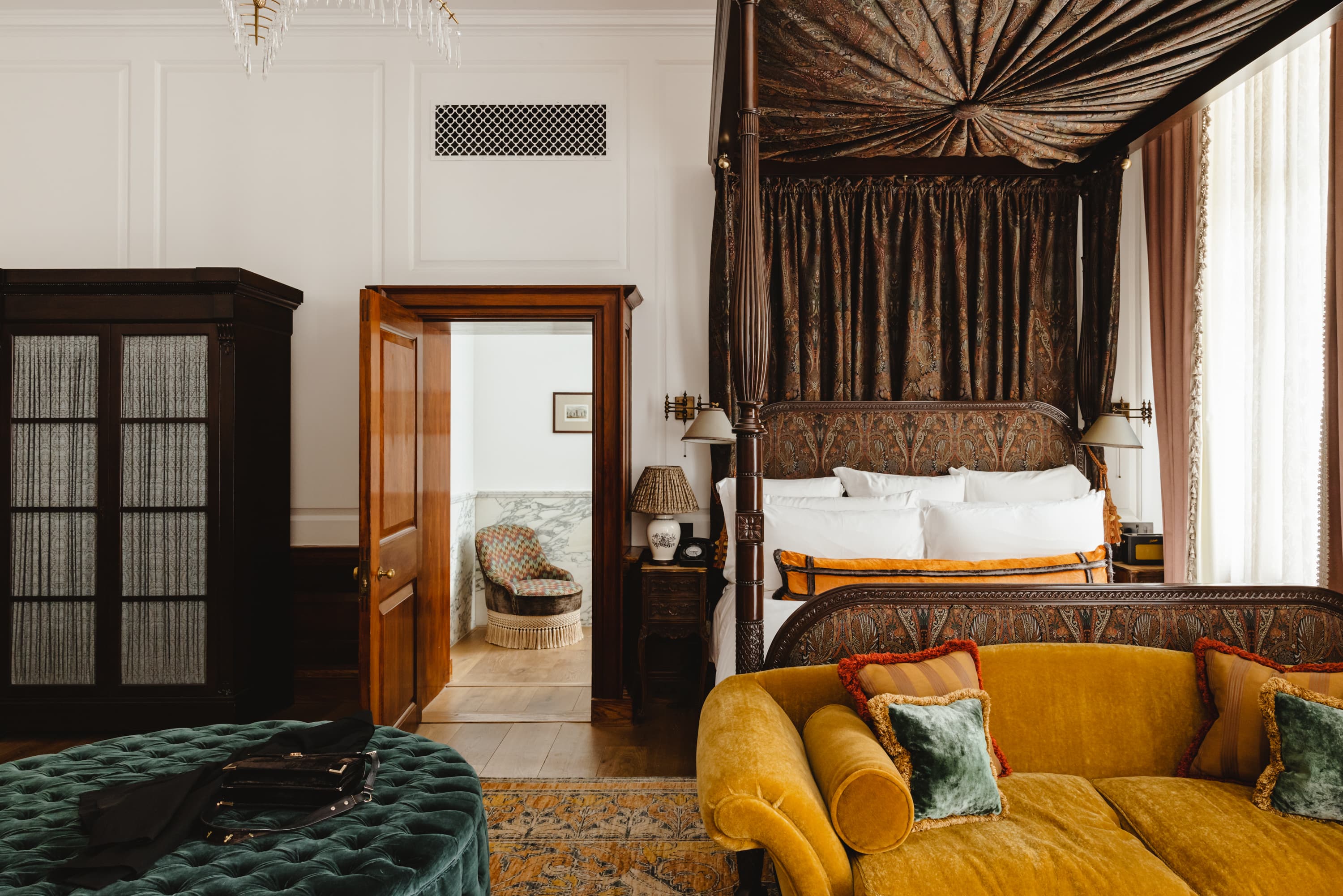 Interior of hotel room at The Ned City of London with plush, velvet furniture and a brown four poster bed.