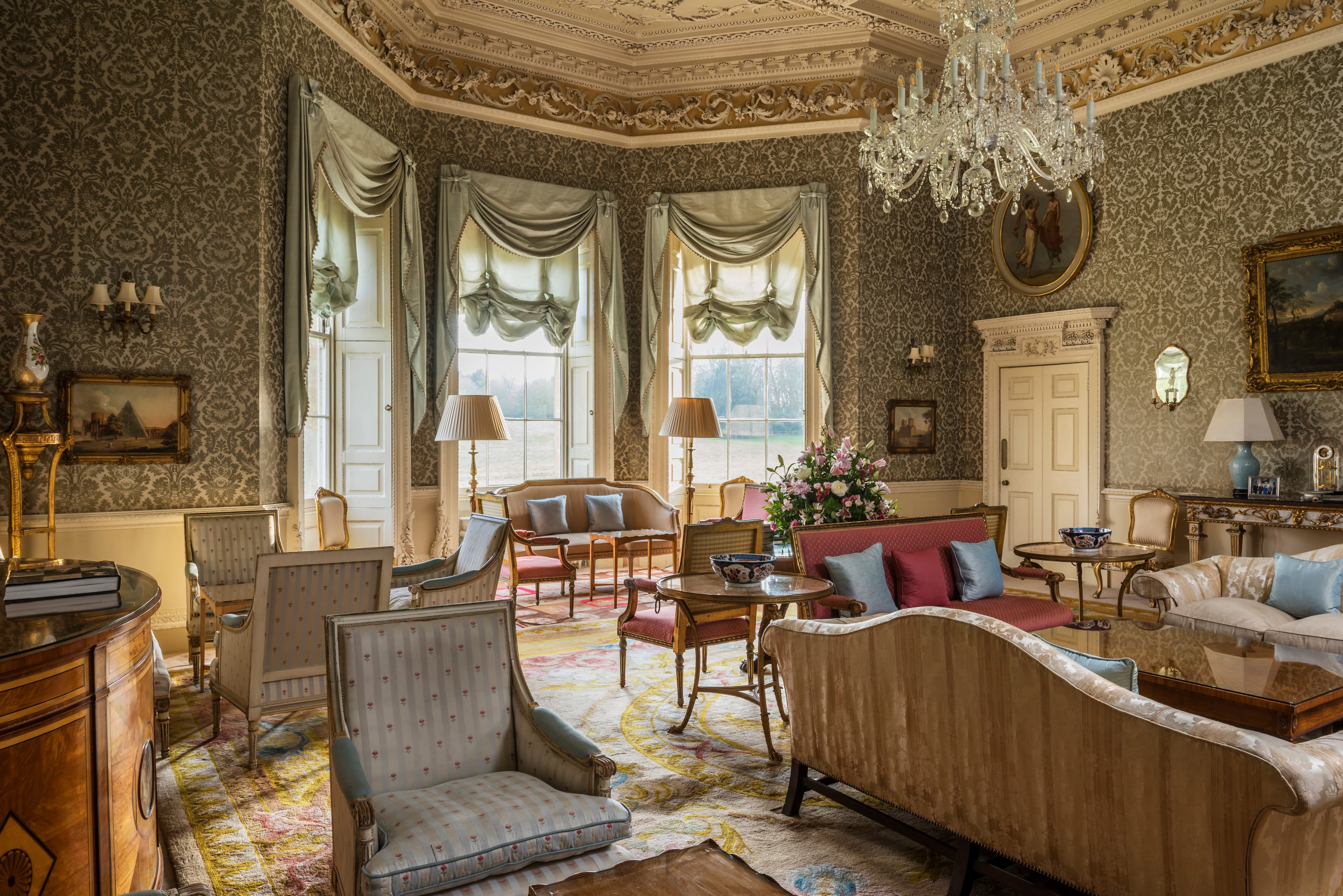 A grand and spacious living room with antique furniture and patterned wallpaper, with a chandelier hanging and embellished ceiling.