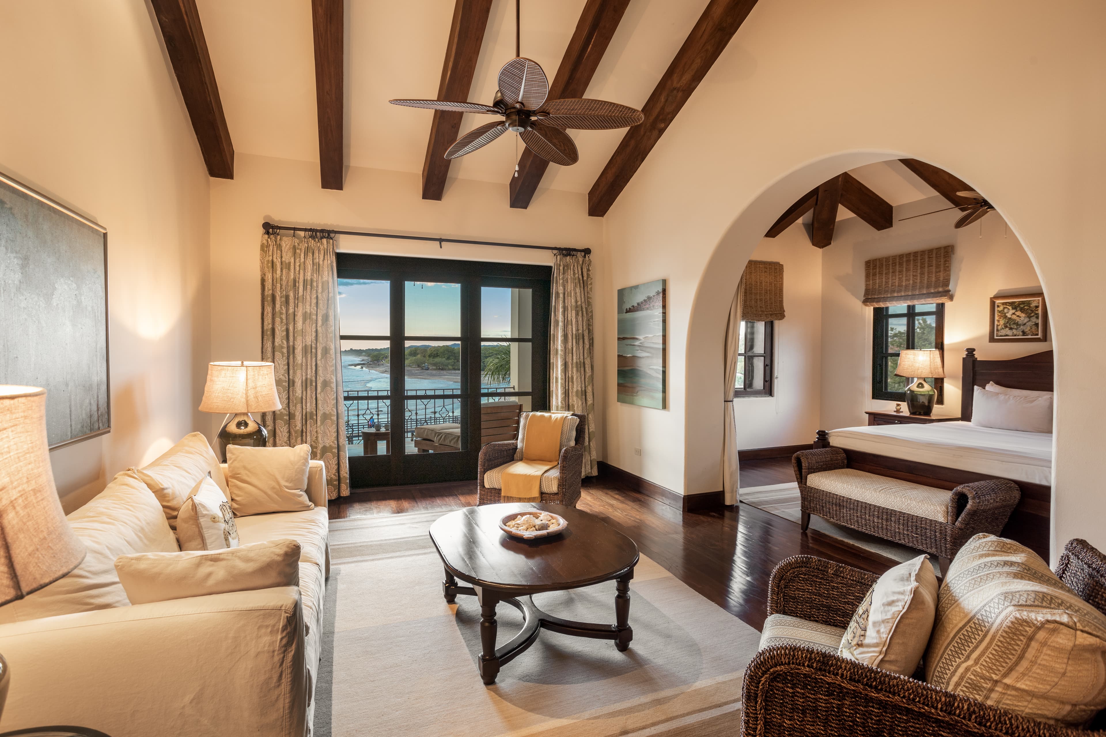 A spacious hotel room with a living room area and bedroom connected by an open arch with wooden beams and furnishings and neutral tones.