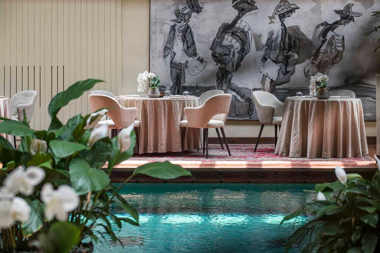 View through plants of a swimming pool with round dining tables set up to one side with tablecloths and a large piece of artwork on the wall behind.