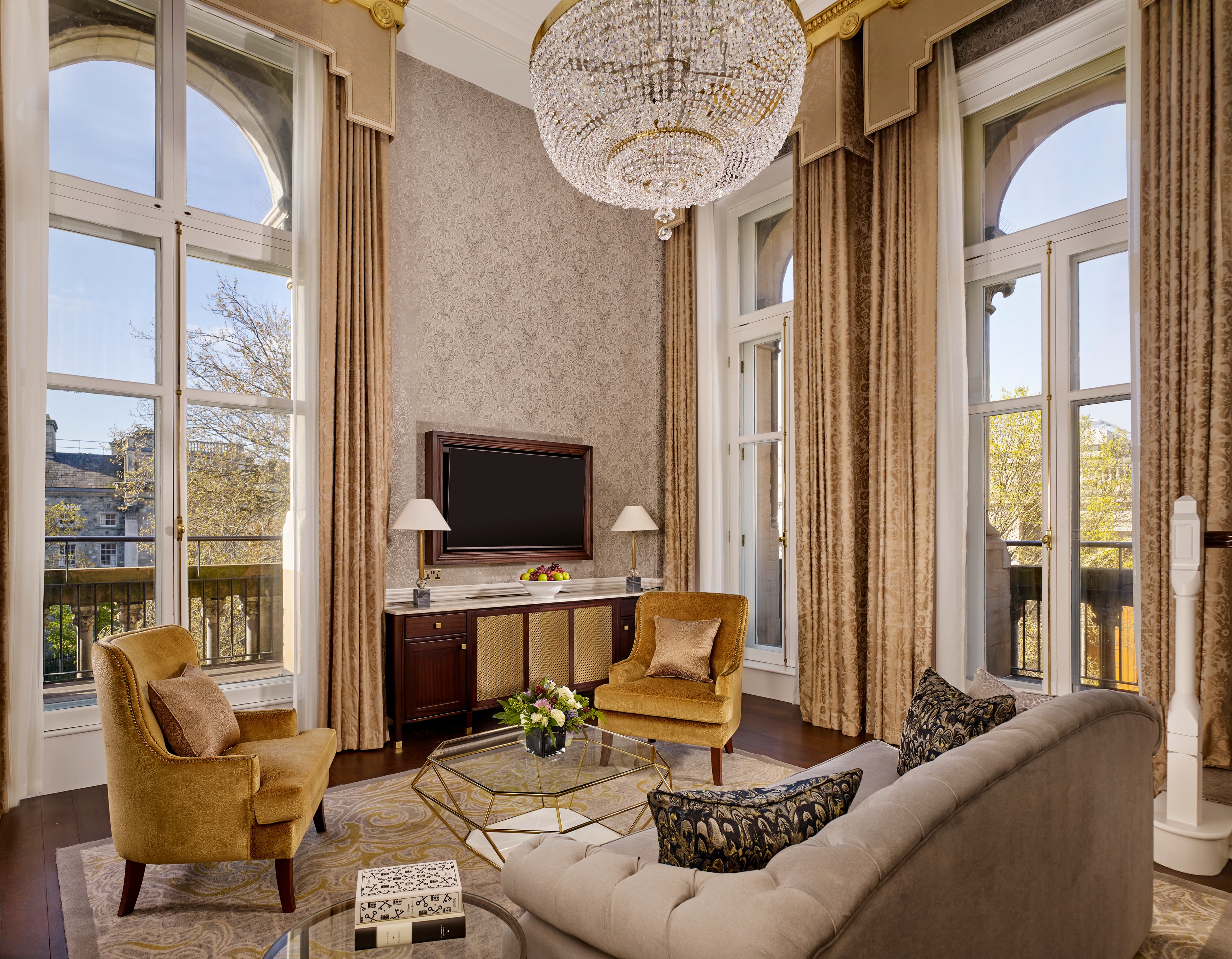 An elegant lounge area of a suite with plush mustard and cream tones, long draped curtains and a chandelier.