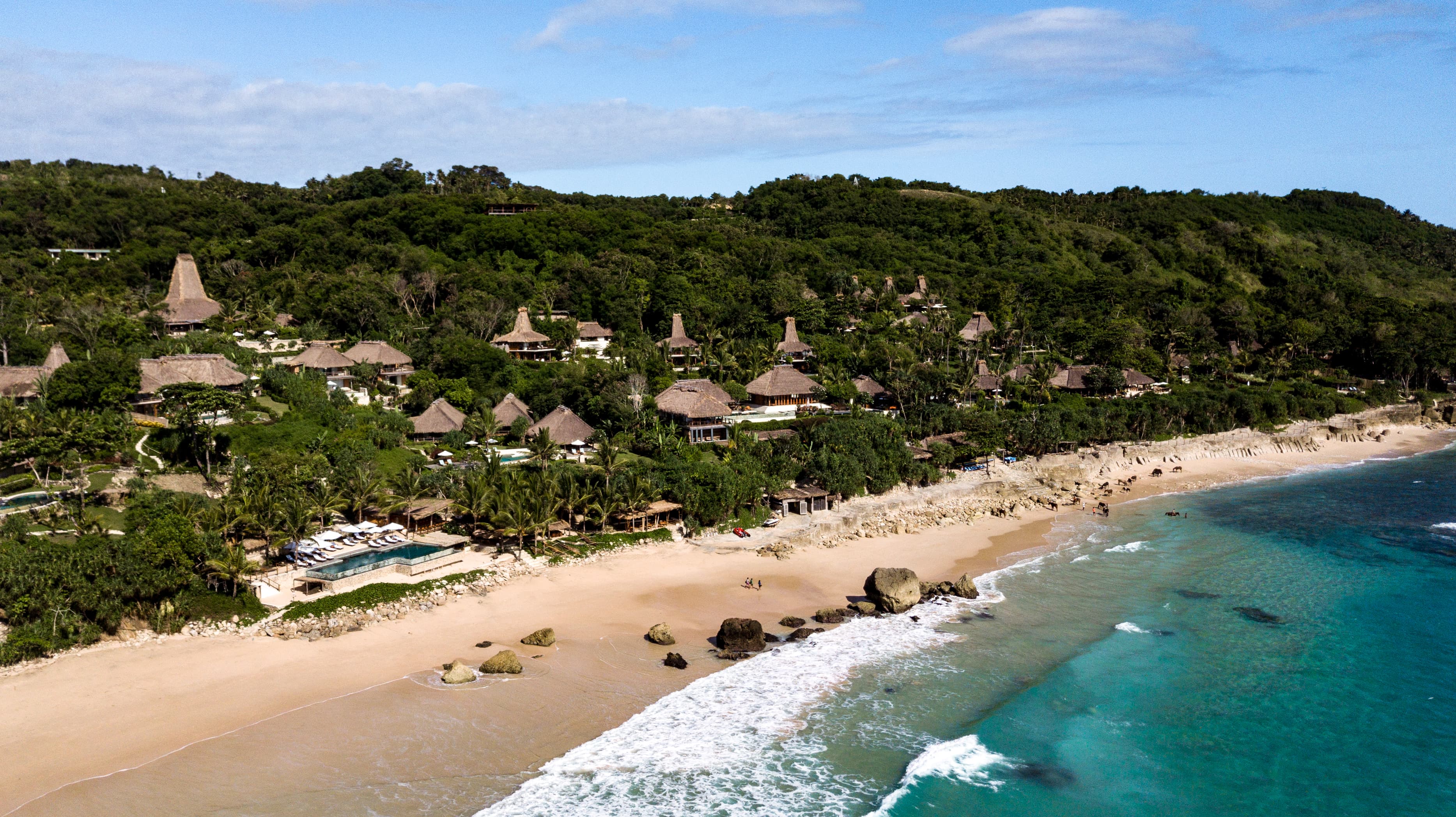 A view of a beutiful beach with a resort nestled in the lush jungle with multiple small huts with straw roofs.
