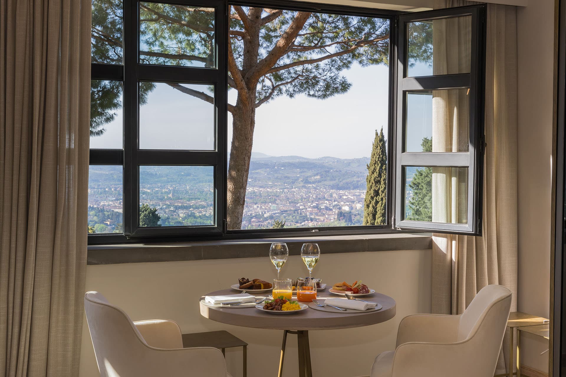 A view of a dining set up with two glasses of wine and food with a large open window looking out onto a countryside view.