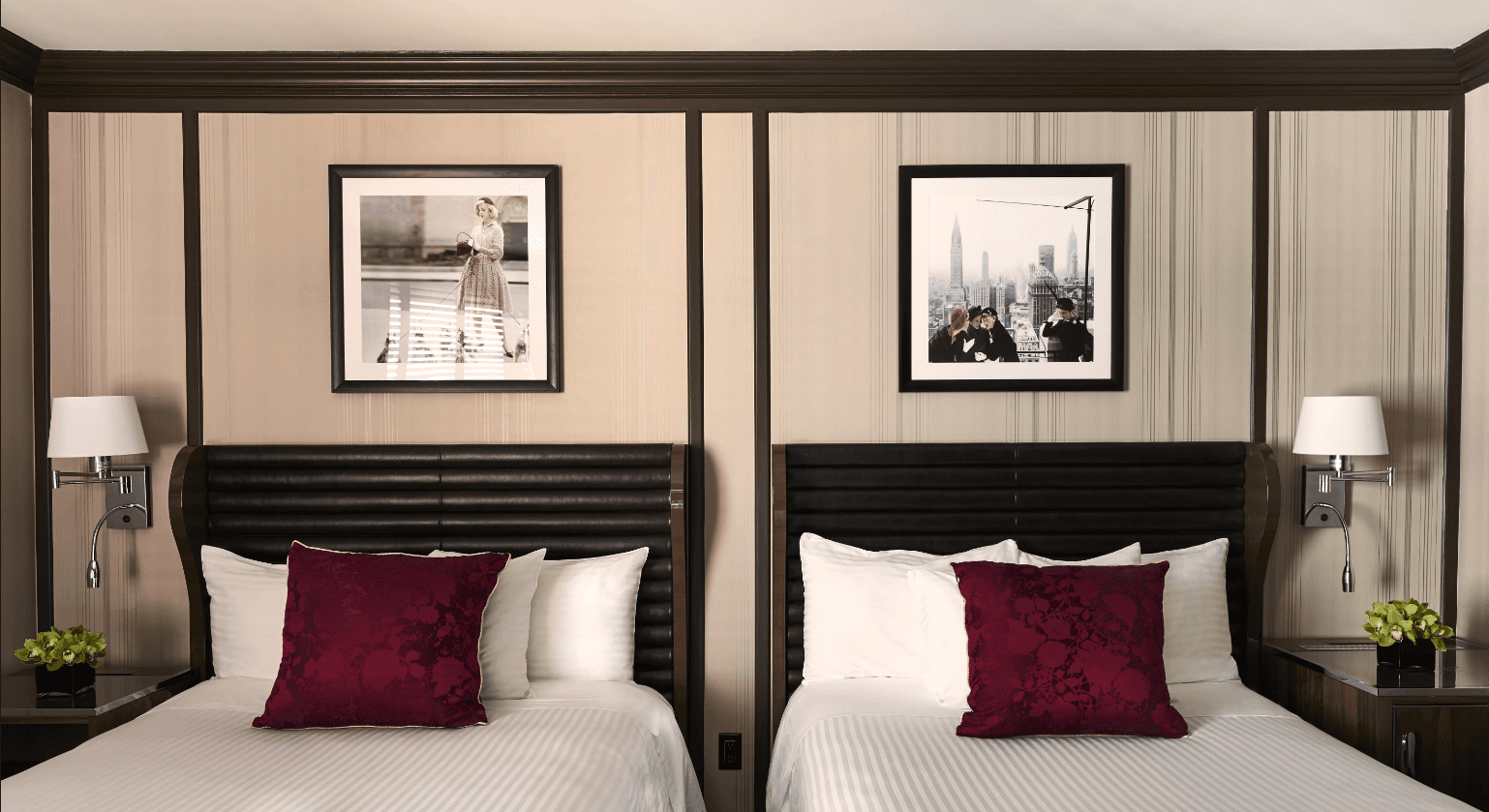 A view of a hotel room at Triumph hotels in NYC with two beds with white bedding and purple cushion and images of NYC on the wall above.