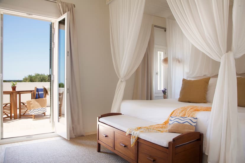 A hotel room with a four poster bed with white fabric and an open door looking out to a balcony with ocean views.