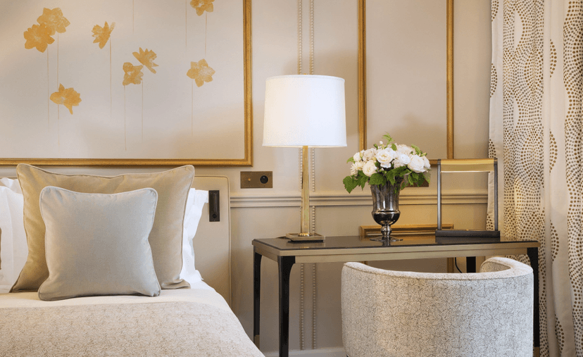 This image shows an elegant hotel room with bed and table and armchair with cream and gold finishes.