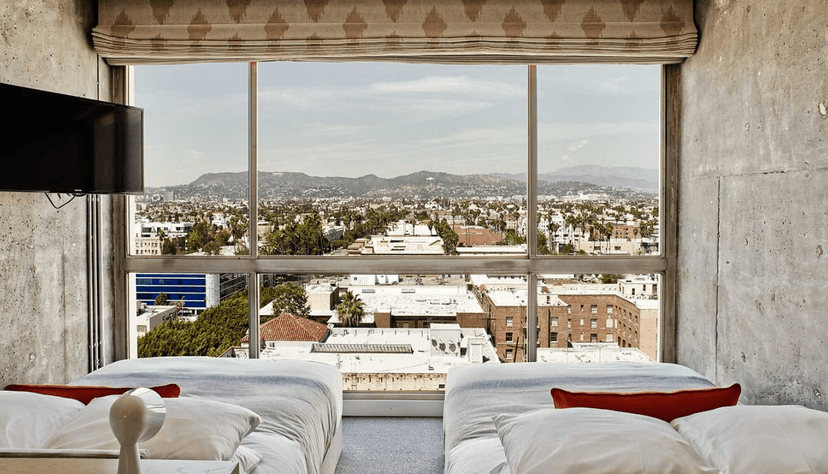 A view of two hotel beds with a large floor-to-ceiling window with a city view.