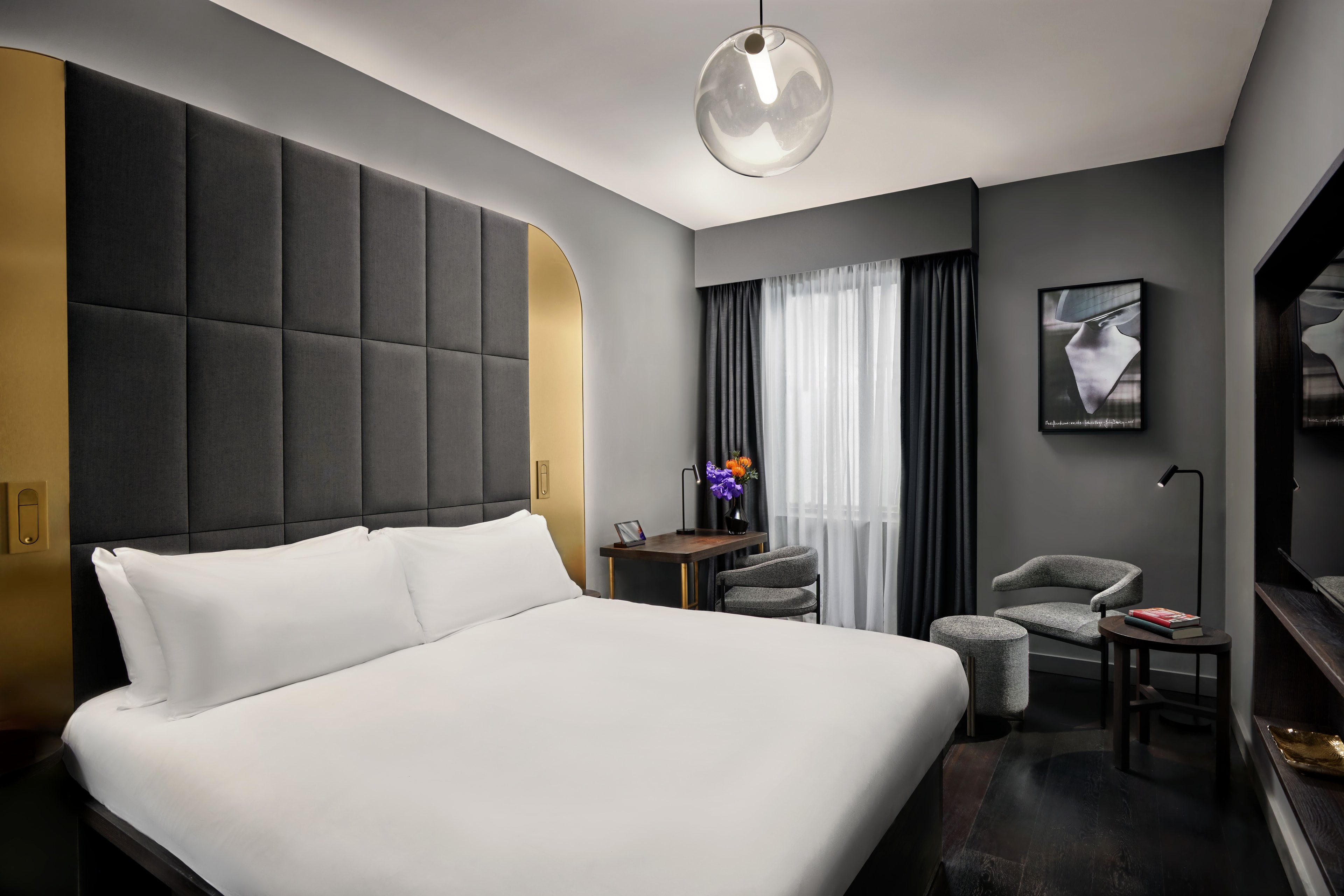 A gray hotel room with a white bed and gray-and-gold headboard with flowers on the nightstand.