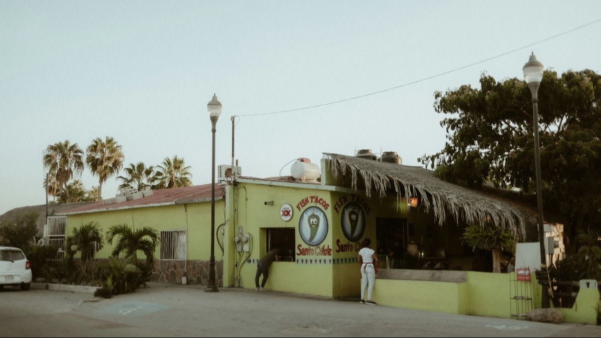 Light green restaurant with a Fish Tacos sign and a section with a straw roof, and palm trees in view