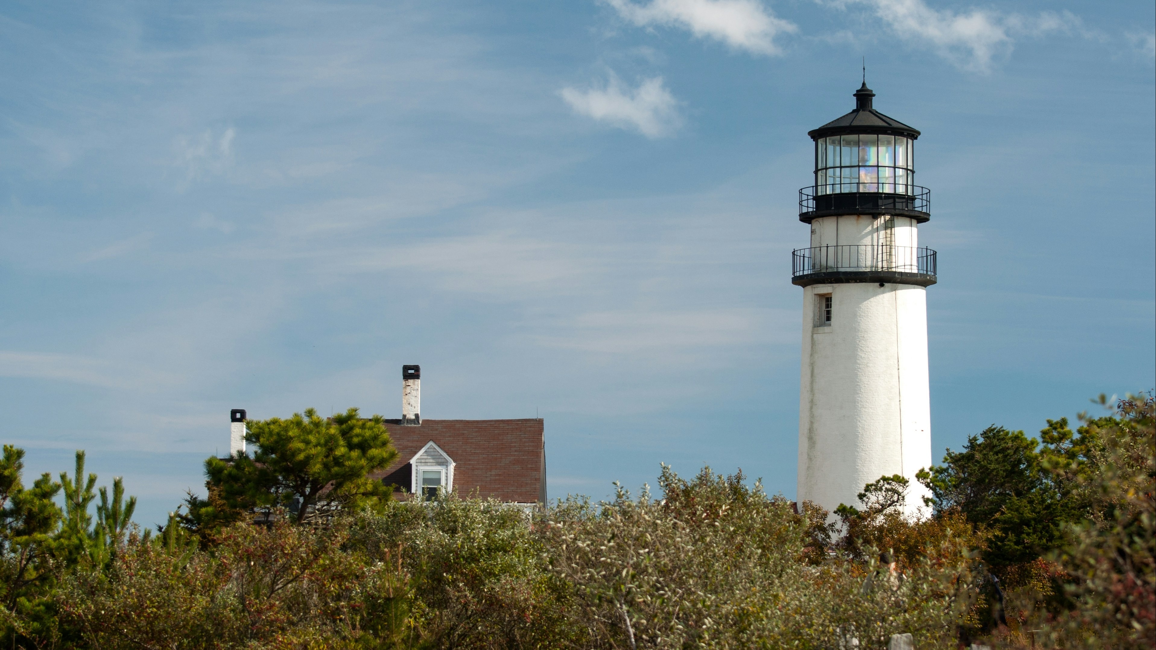 A white lighthouse next to a house in a field on a sunny day in Cape Cod.
