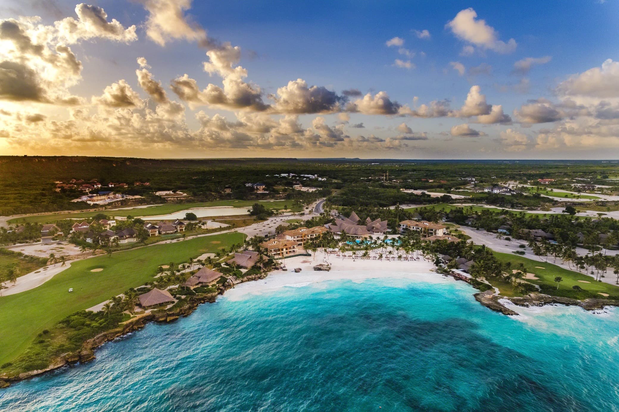 aerial shot of a resort on a white beach with turquoise waters and a sunset behind