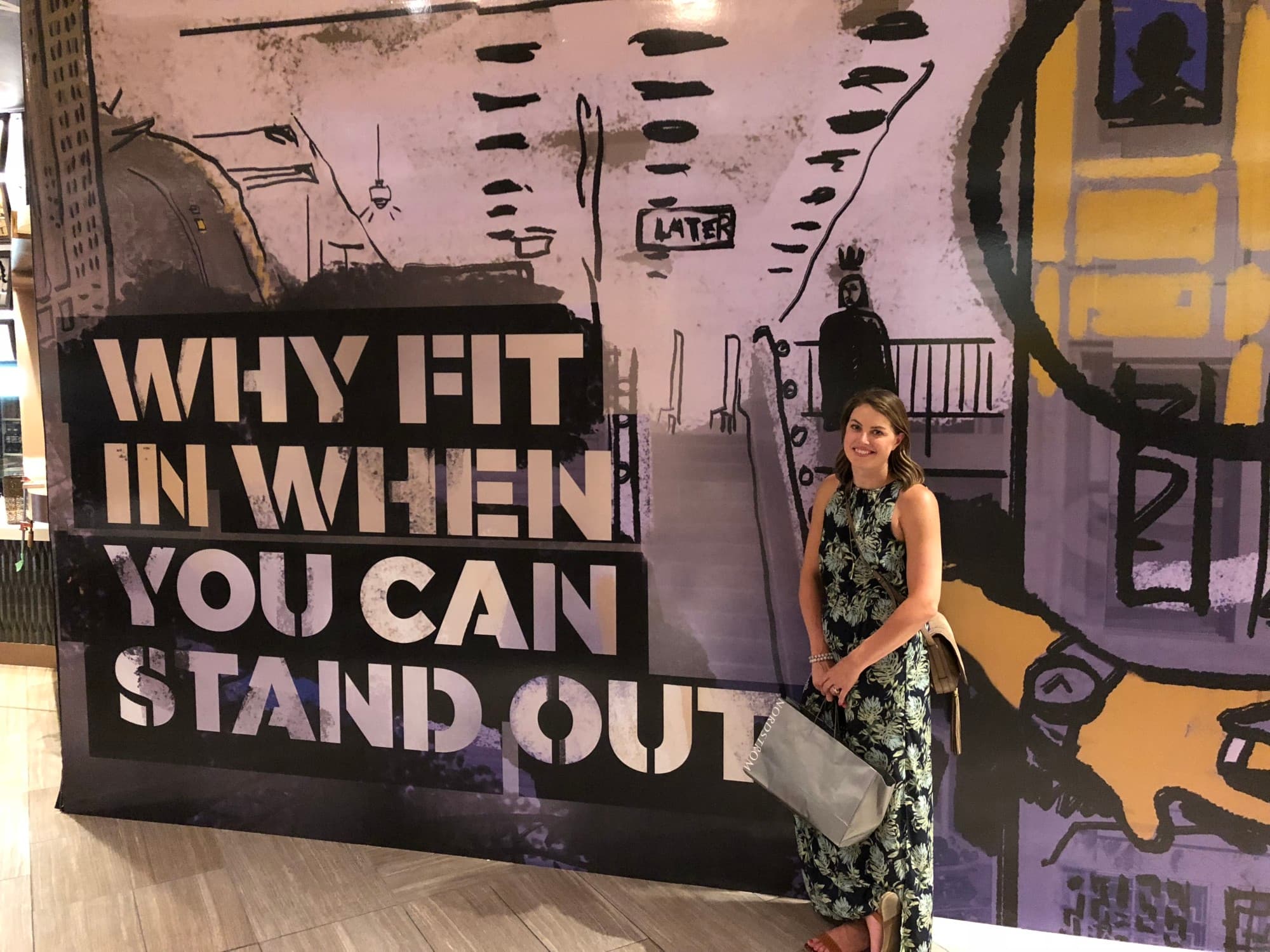 A woman with a shopping bag stands in front of a mural that says, "Why fit in when you can stand out?"