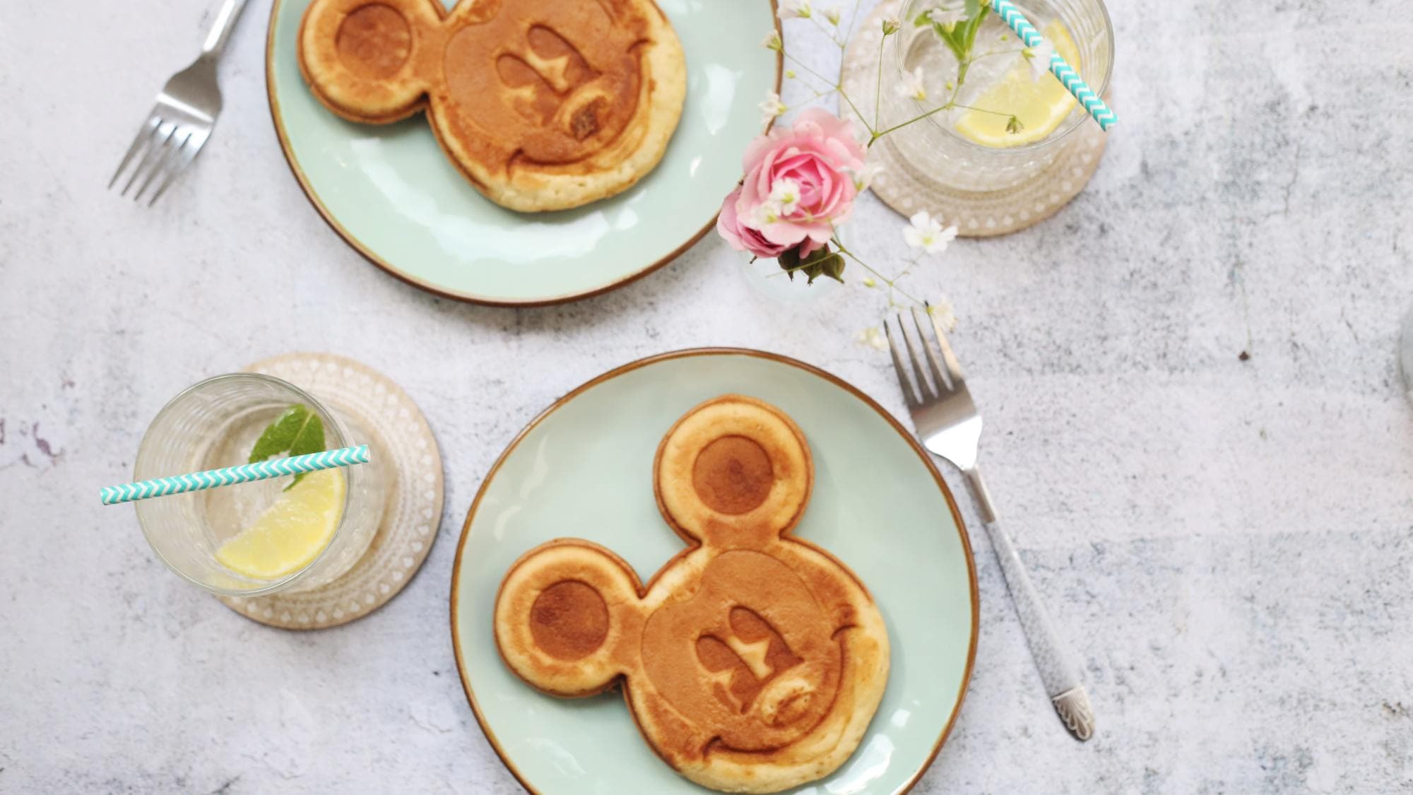 Mickey Mouse shapes pancakes on delicate pastel dishes along with flowers and lemon-infused water.