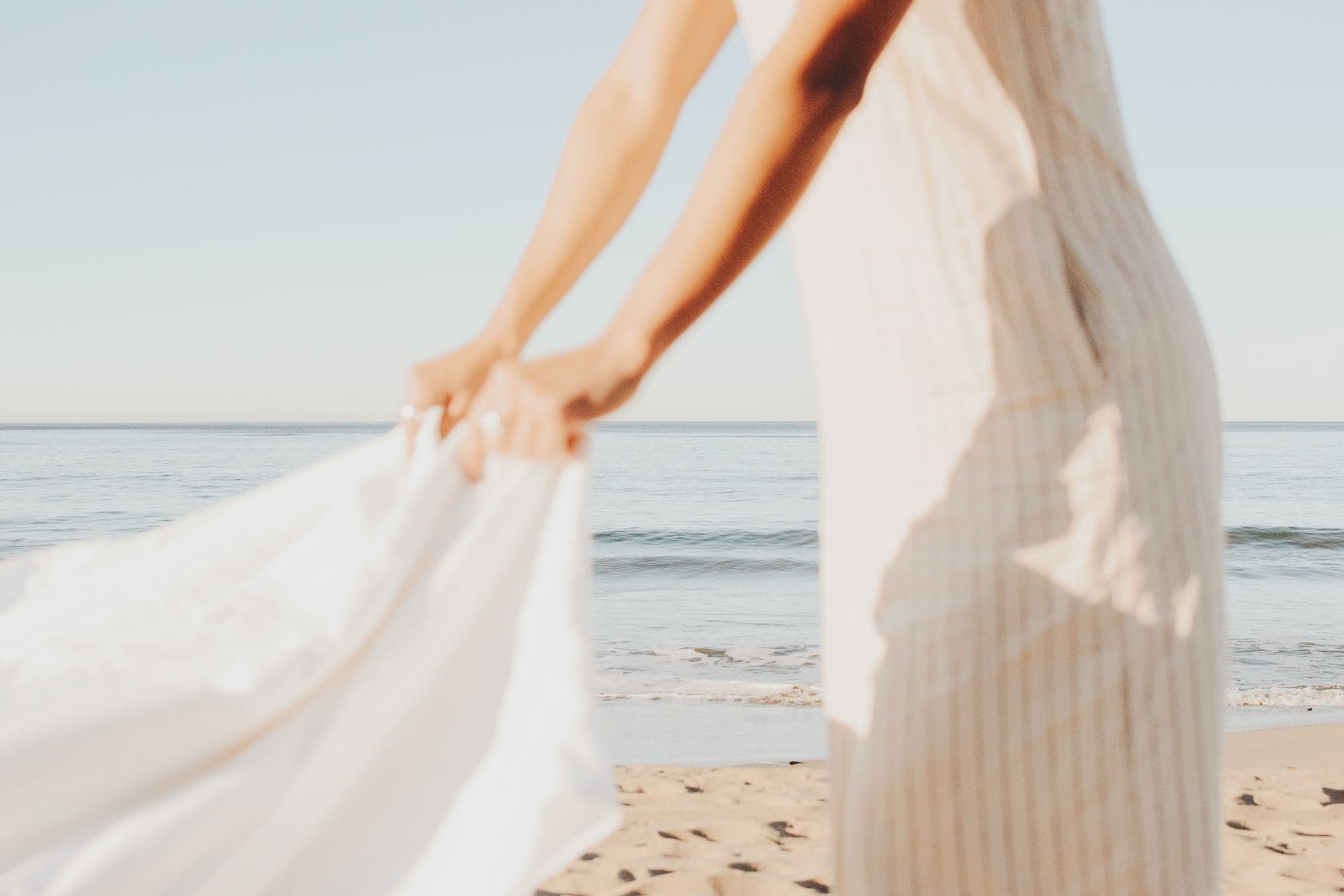 a woman in a linen dress lays down a white blanket on a beach