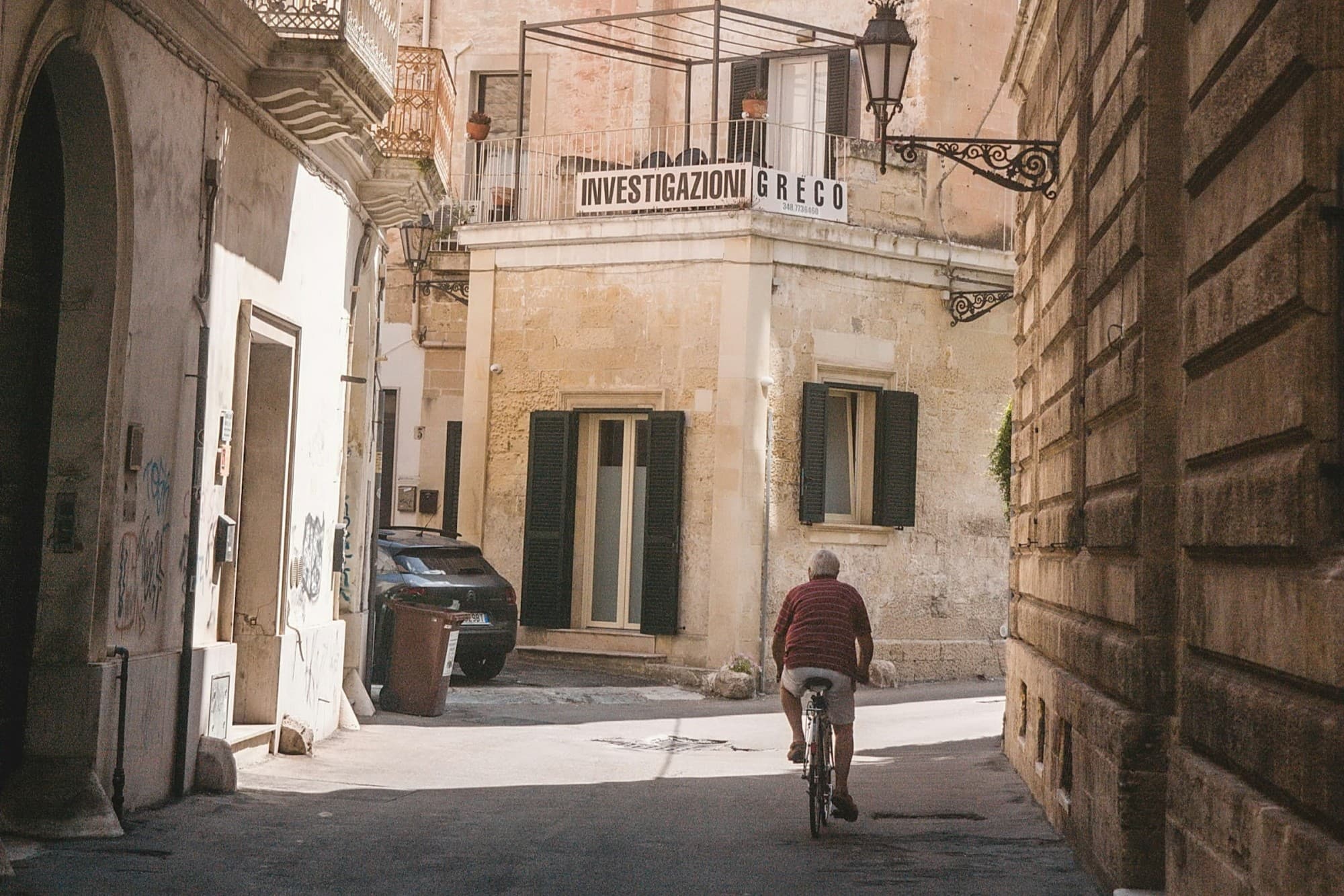 A lone, elderly man bikes along an alleyway in a historic section of Lecce, with a sign that reads "Investigazioni Greco" at the end