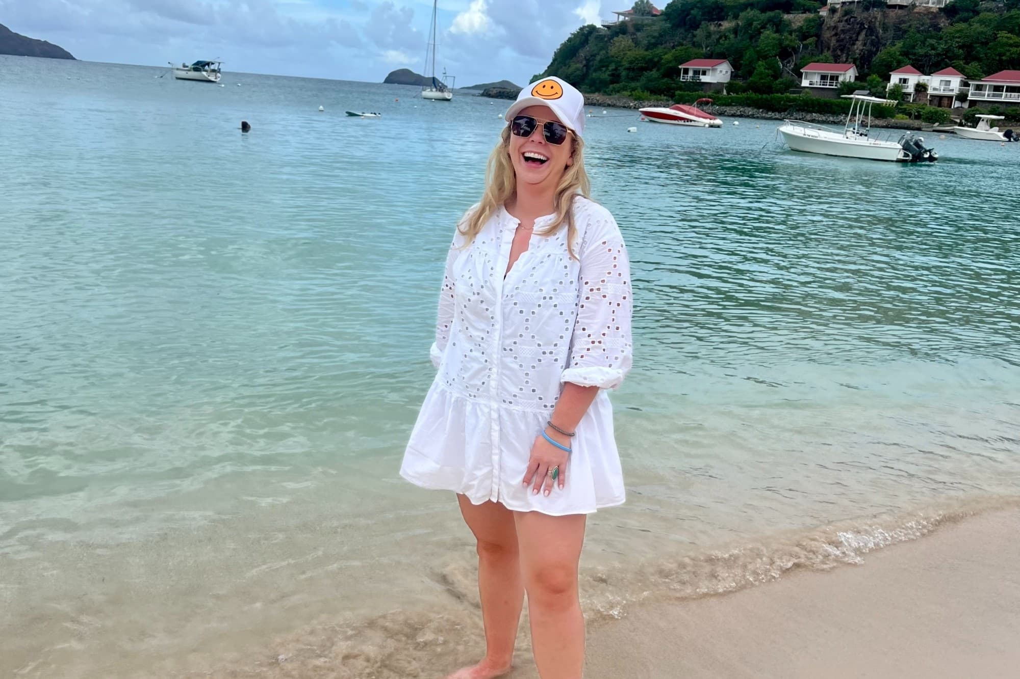 a woman in a white dress and a baseball cap laughs on a beach