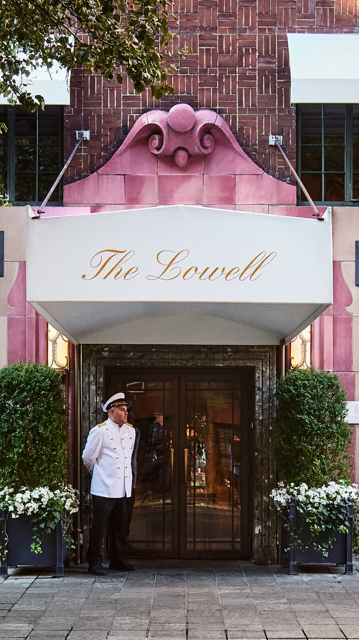 a man in a white coat stands in front of an elegant building façade with an awning that reads "The Lowell."