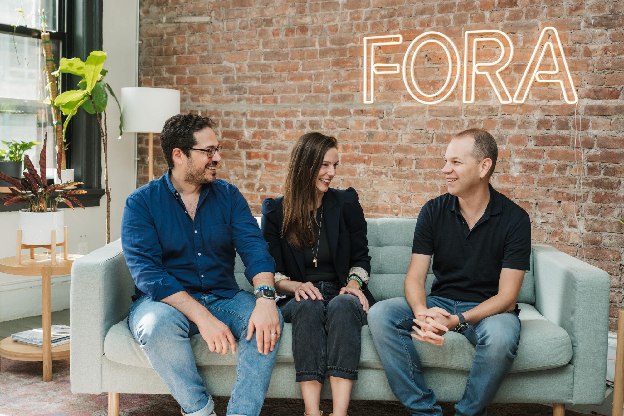 A woman and two men smile at each other on a couch in front of a brick wall