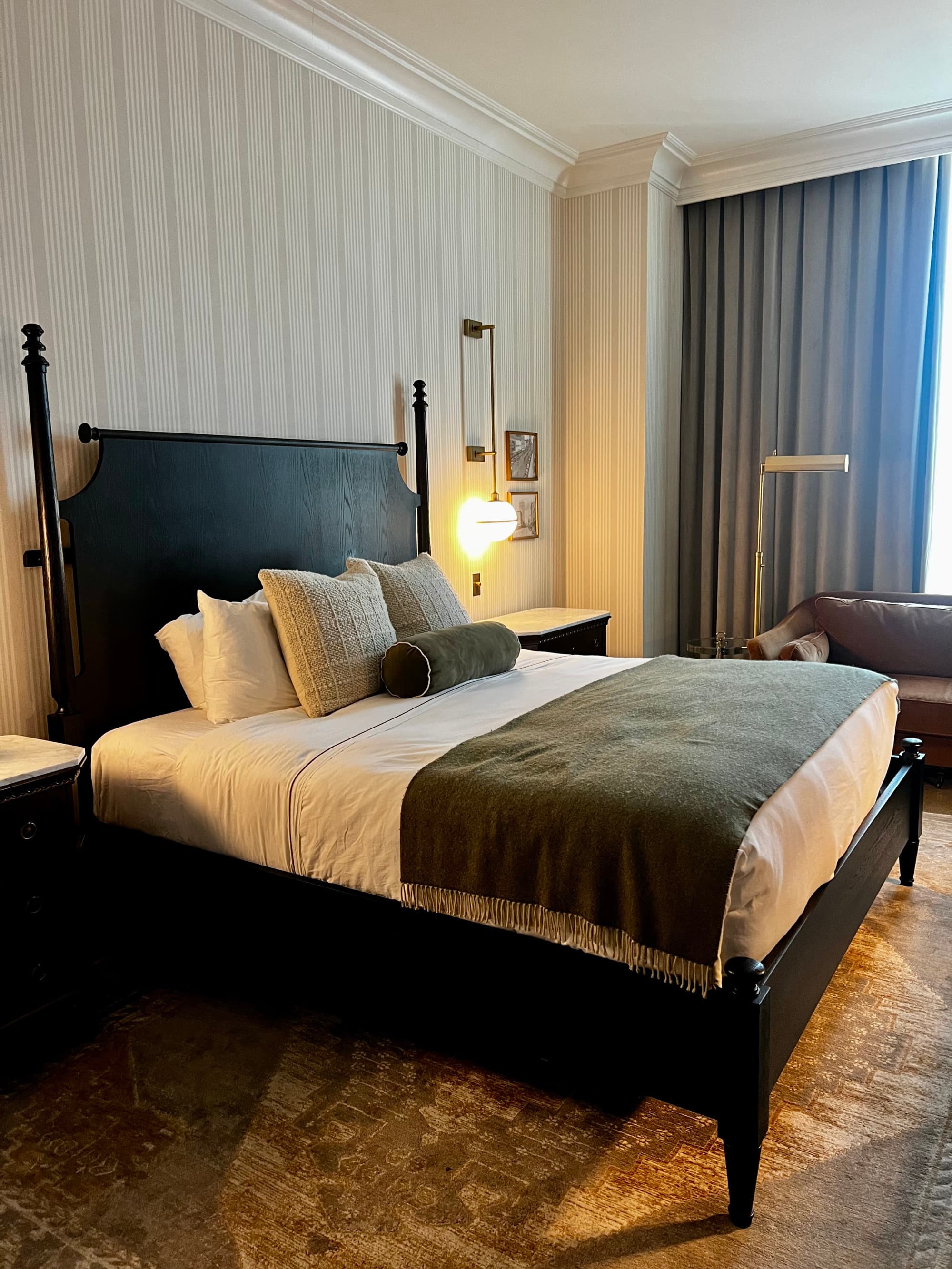 A view inside a hotel room with white pinstriped walls, a dark wood bedframe and headboard with white bedding and dark curtains.